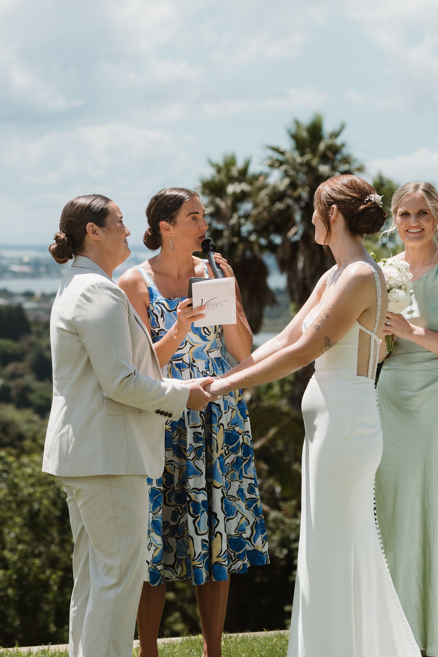 Two brides hold hands during an outdoor wedding ceremony officiated by a woman in a floral dress, with a bridesmaid nearby.