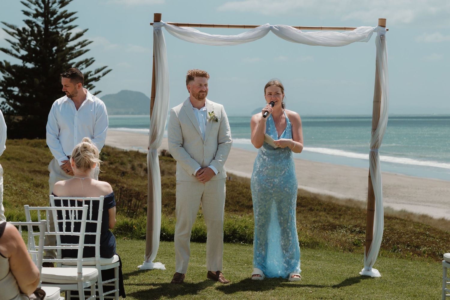 Outdoor beach wedding ceremony with groom in tan suit and officiant in blue sequin dress under a draped wooden arch.