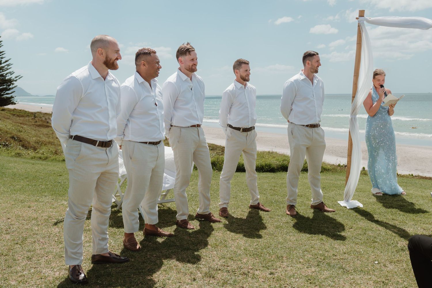 Groomsmen in white shirts and tan pants stand outdoors near beach at wedding ceremony.
