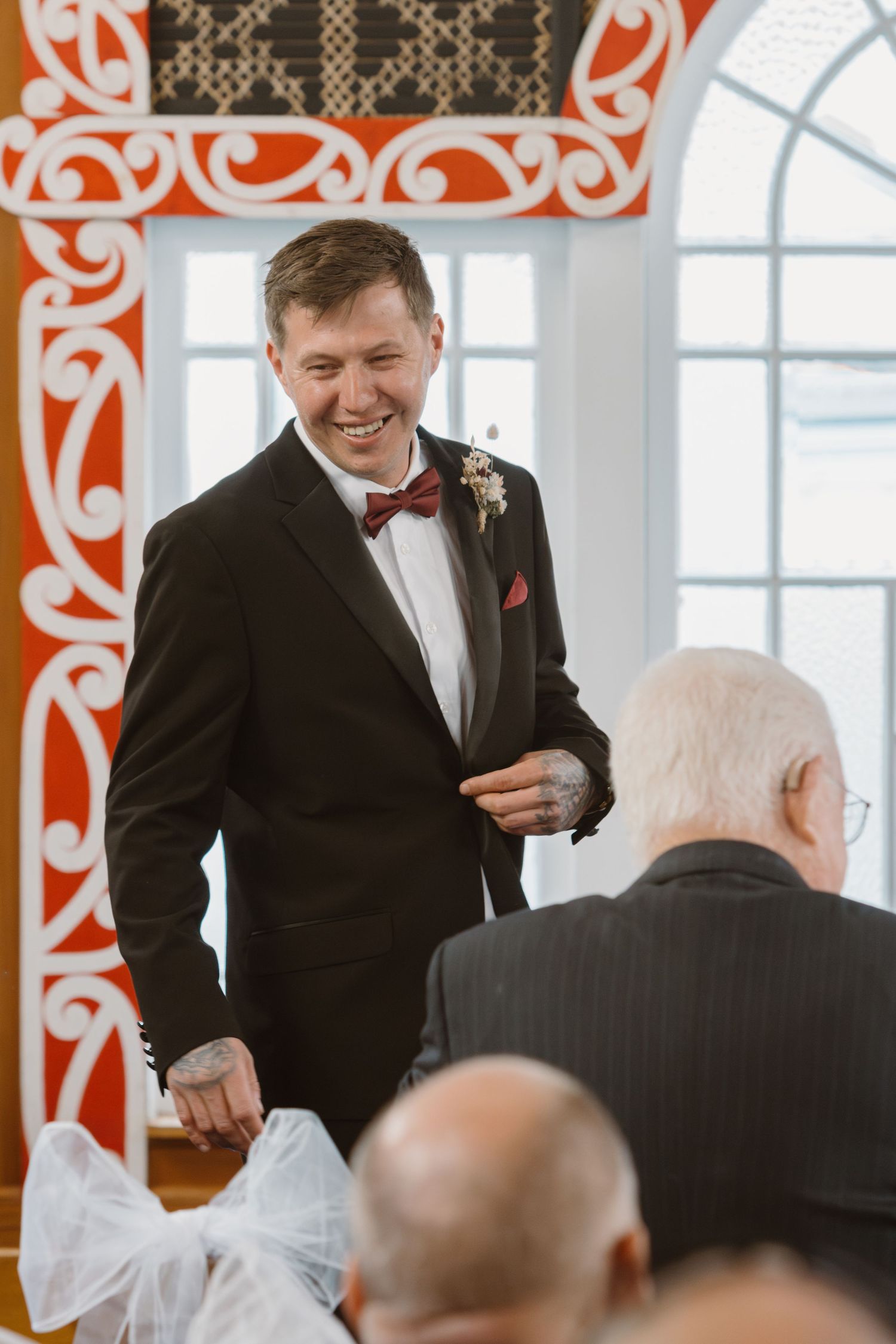 A groom in a black tuxedo and red bow tie laughs joyfully during the wedding reception with guests.