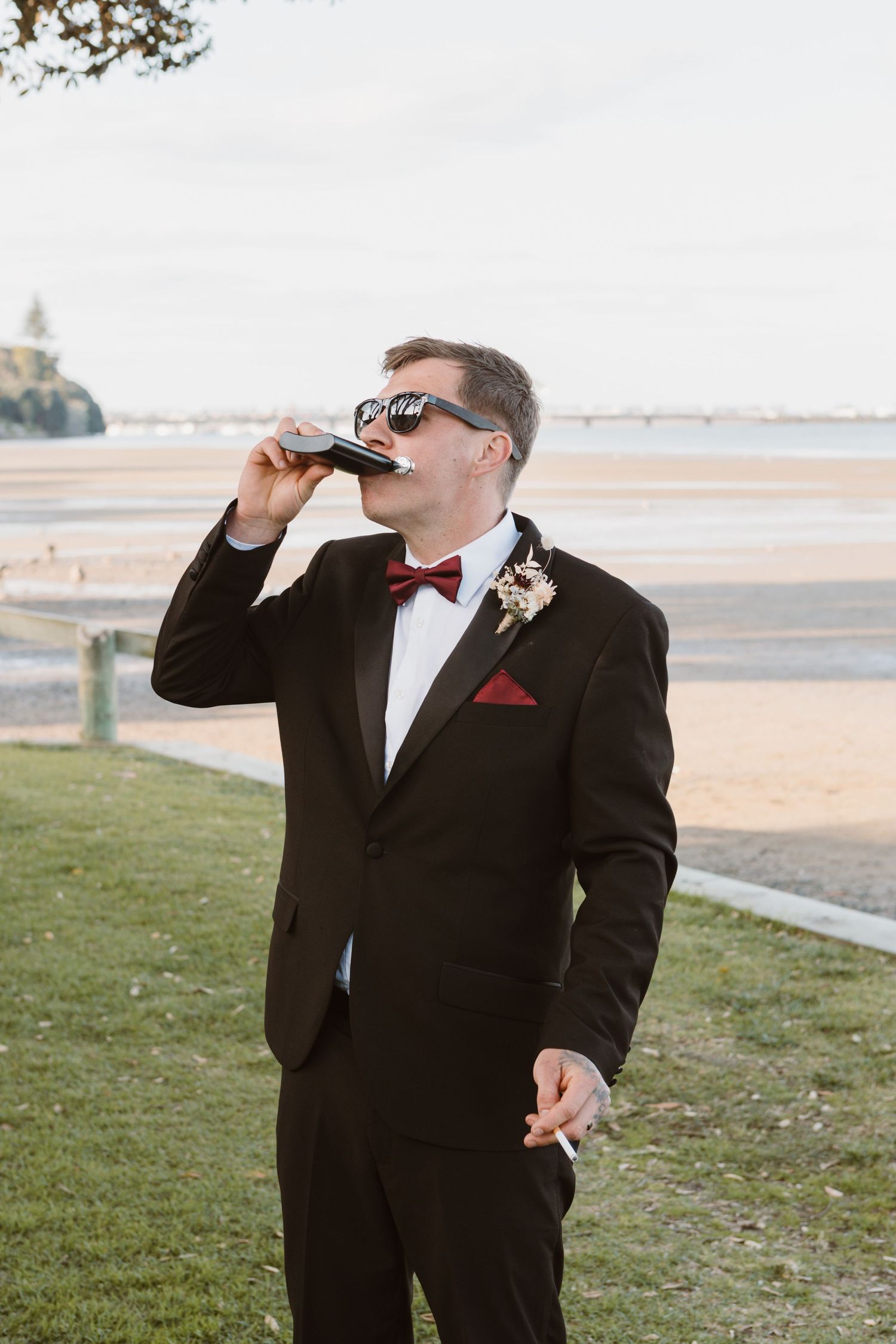 A person in a black tuxedo and red bow tie takes a drink while standing outdoors near a beach.