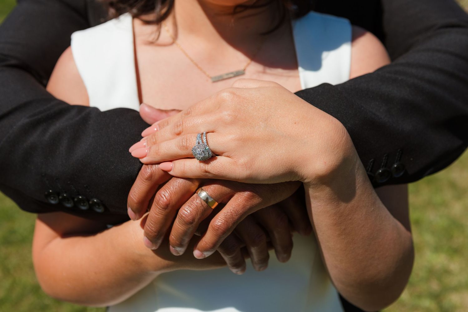 Close-up of hands stacked together showing a sparkling engagement ring against a sunny outdoor background.