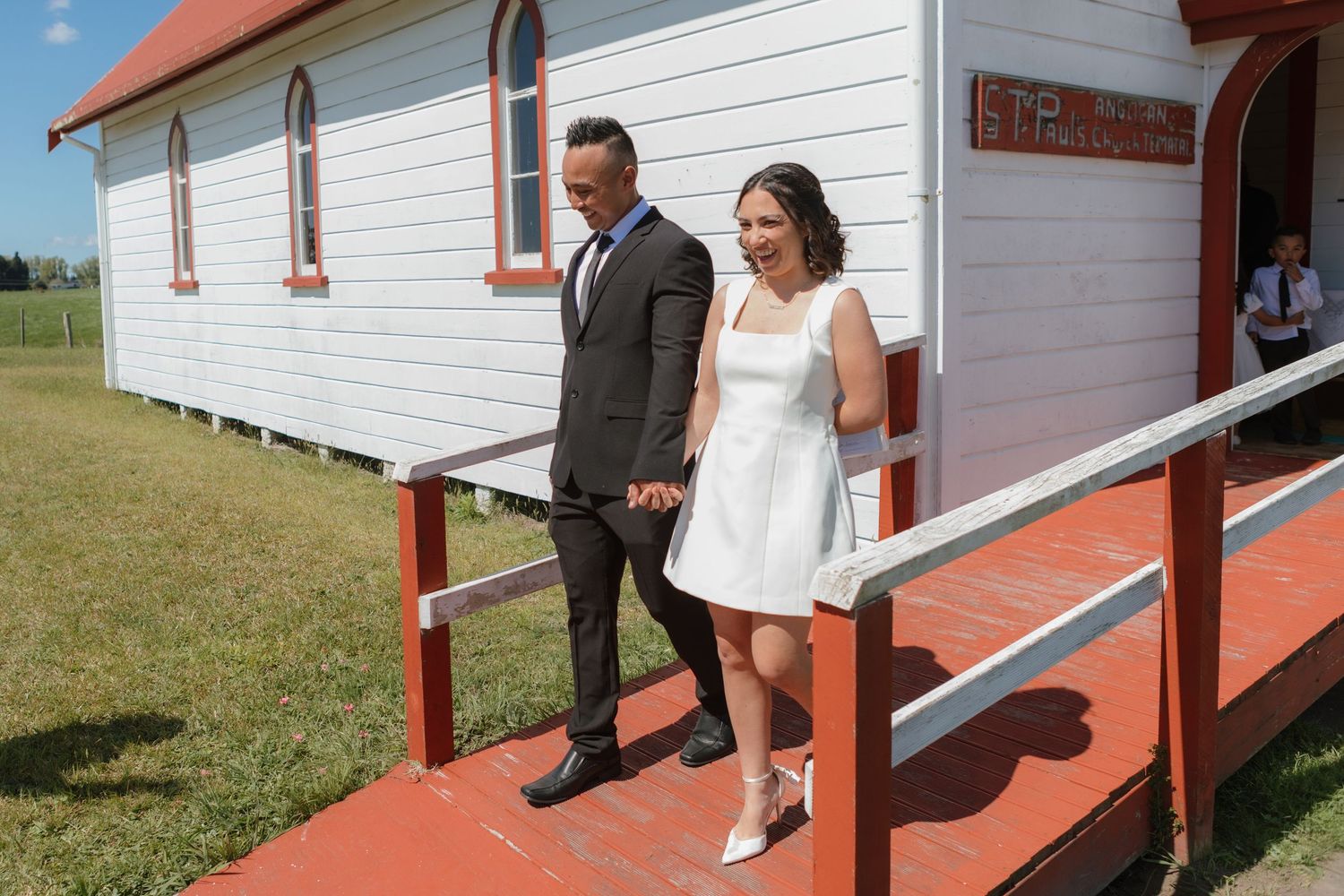 A couple exits a small white church with red trim on a sunny day, with the bride wearing a short white dress.