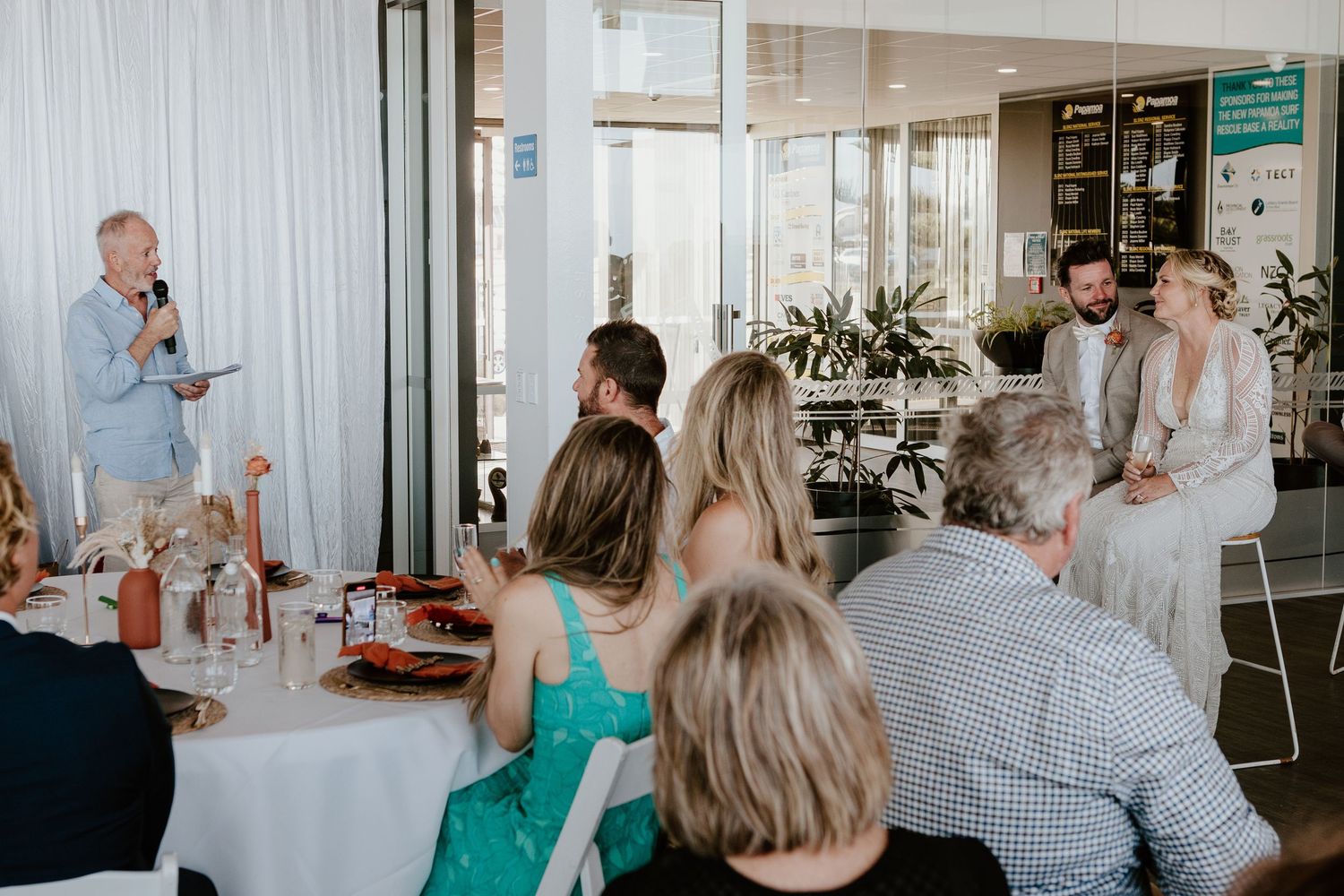 A person delivers a speech at a small wedding reception with guests seated at decorated tables in a bright venue.