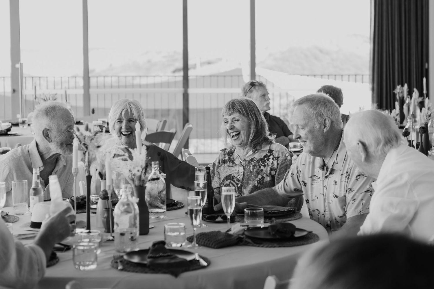 A black and white wedding reception scene shows guests dining at decorated tables with a scenic outdoor view.