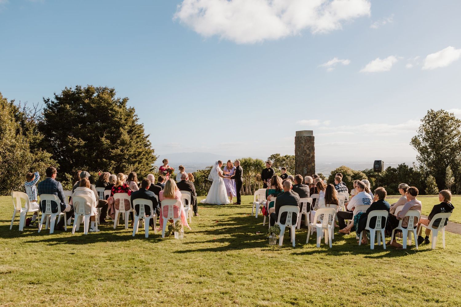 An outdoor wedding ceremony takes place on a hilltop lawn with guests seated in white chairs under a blue summer sky.