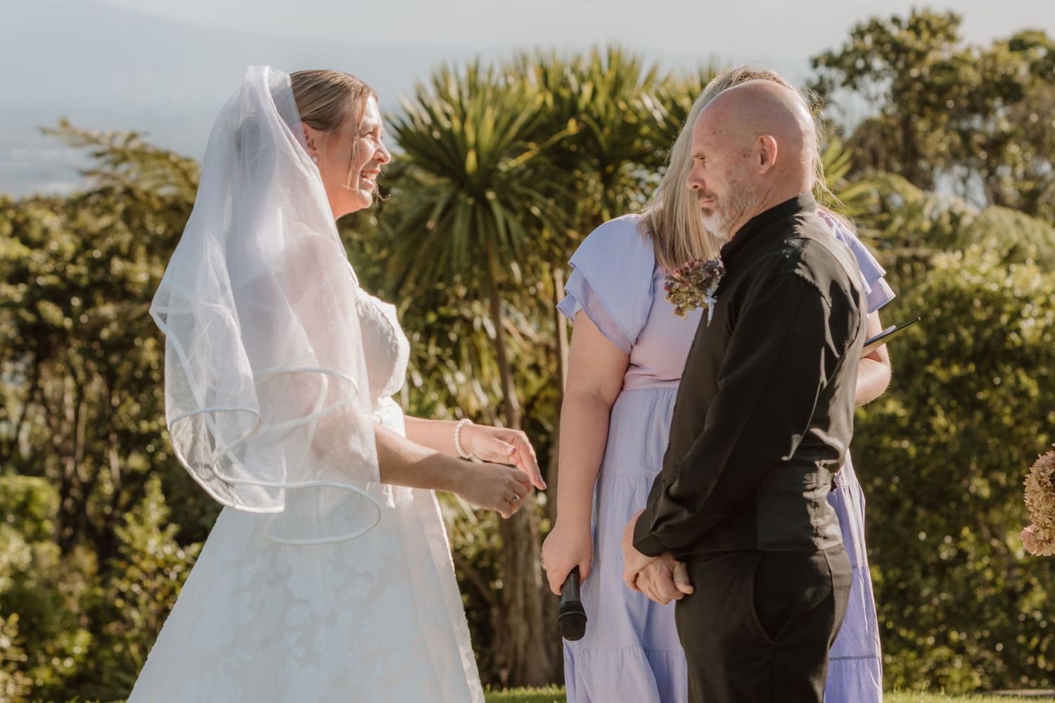 A wedding ceremony takes place in a lush outdoor garden setting with palm trees and natural greenery in the background.