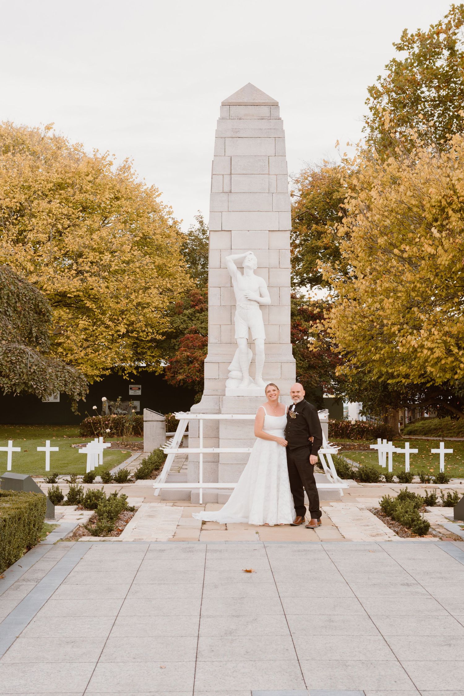 A romantic wedding photoshoot series at a memorial monument featuring white crosses and fall foliage in the background.