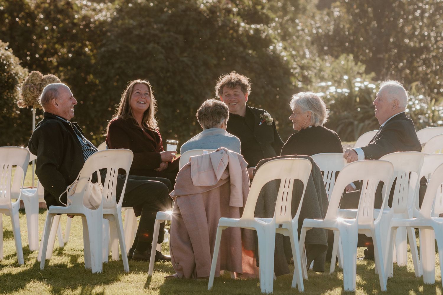 People seated in white chairs outdoors during a sunlit golden hour event with trees in the background.