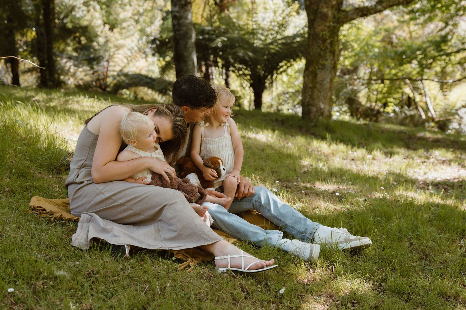 Family sitting together on grass in a sunny park, parents bonding with young children outdoors on a warm day.