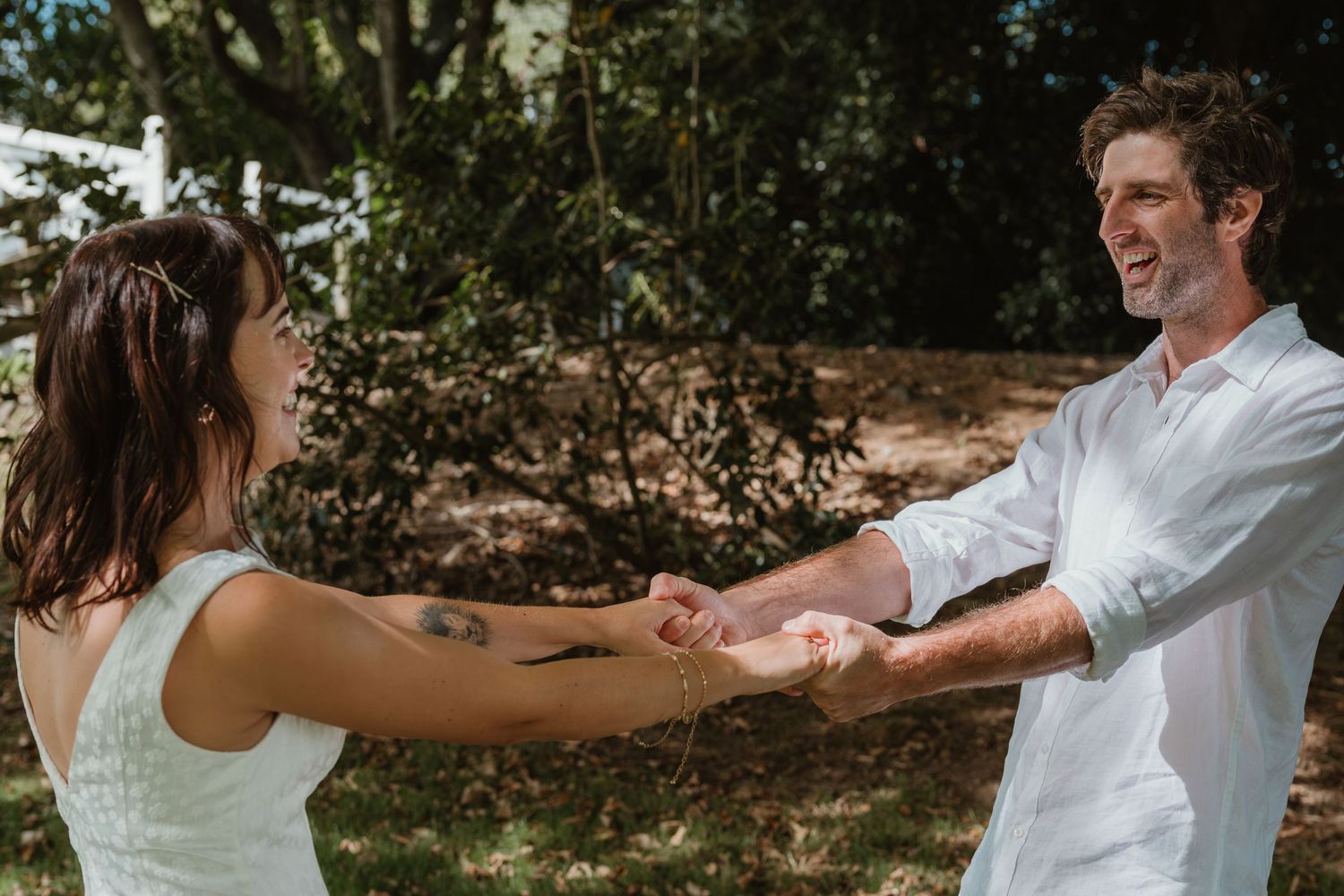 A joyful couple holds hands outdoors, smiling at each other in a sunlit garden setting.