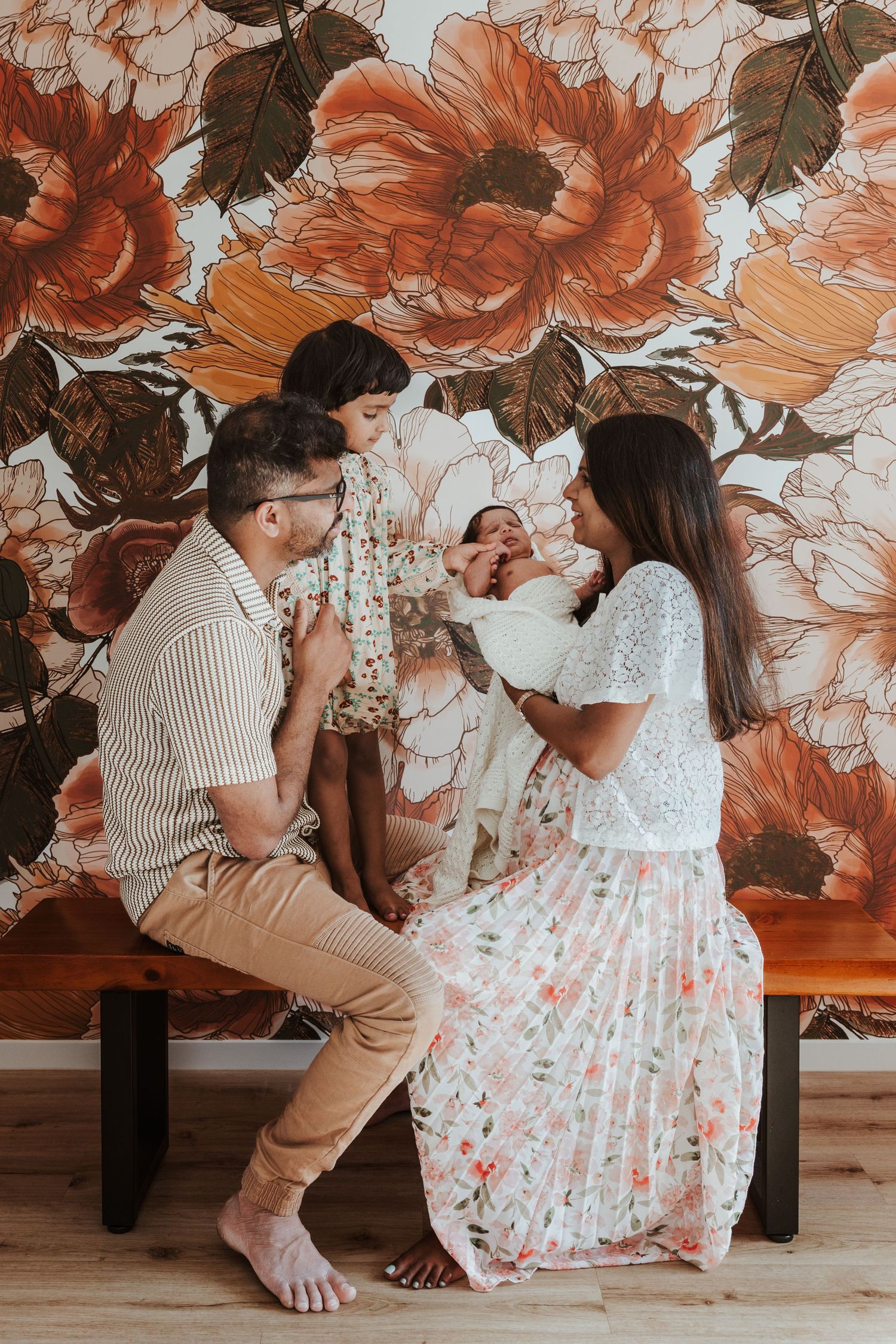 Two people in casual attire sit together on a wooden floor against a floral wallpaper backdrop in warm orange tones.