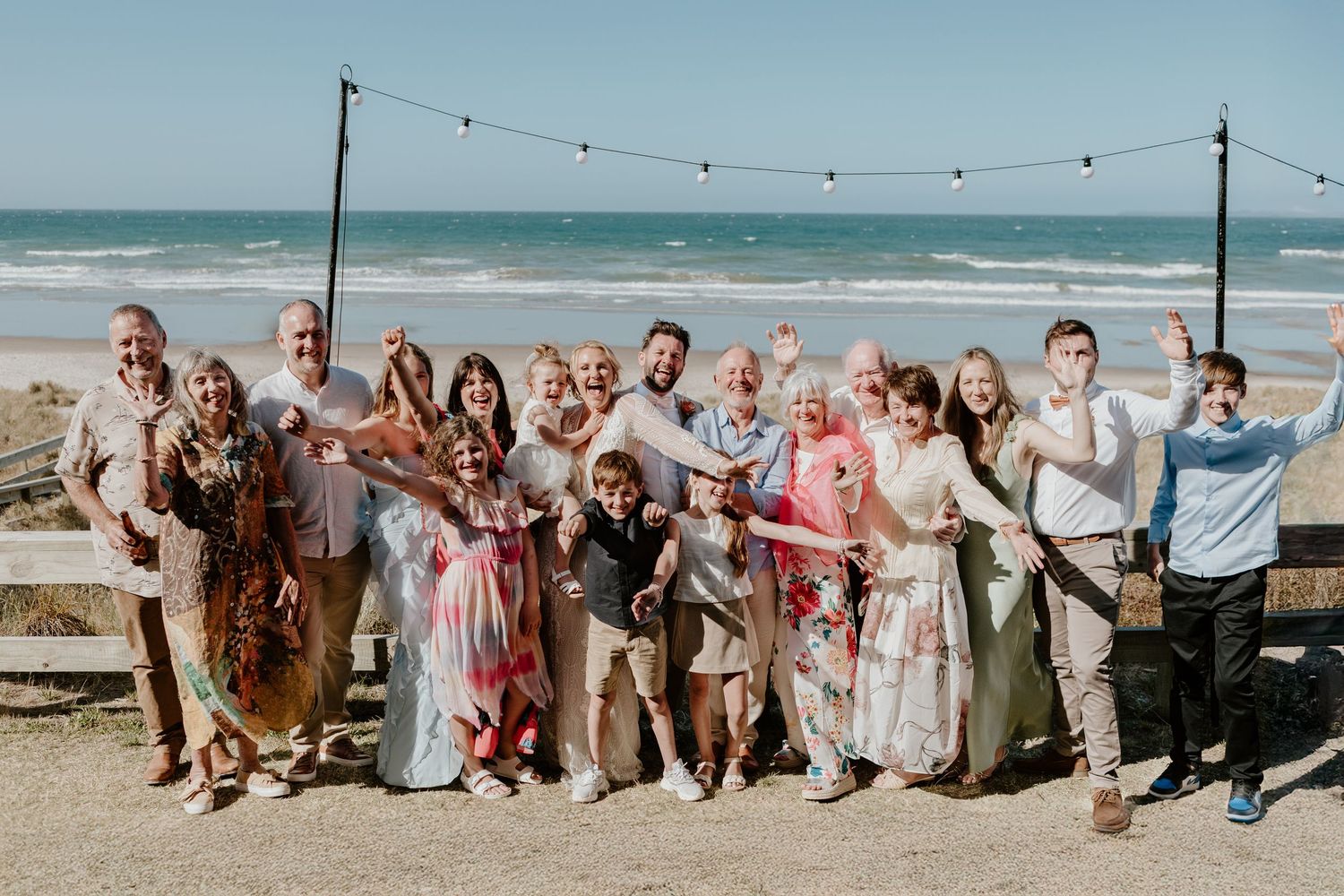 A joyful gathering of people celebrating on a sunny beach with string lights and ocean waves in the background.