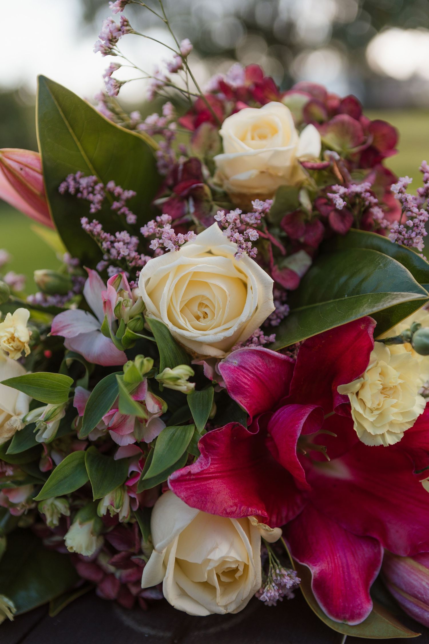 Close-up of elegant floral arrangement with cream roses, deep red lilies, and delicate purple flowers with green foliage.