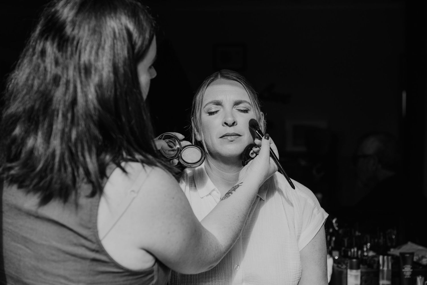 A black and white photograph shows someone applying makeup to another person's face in a darkened setting.