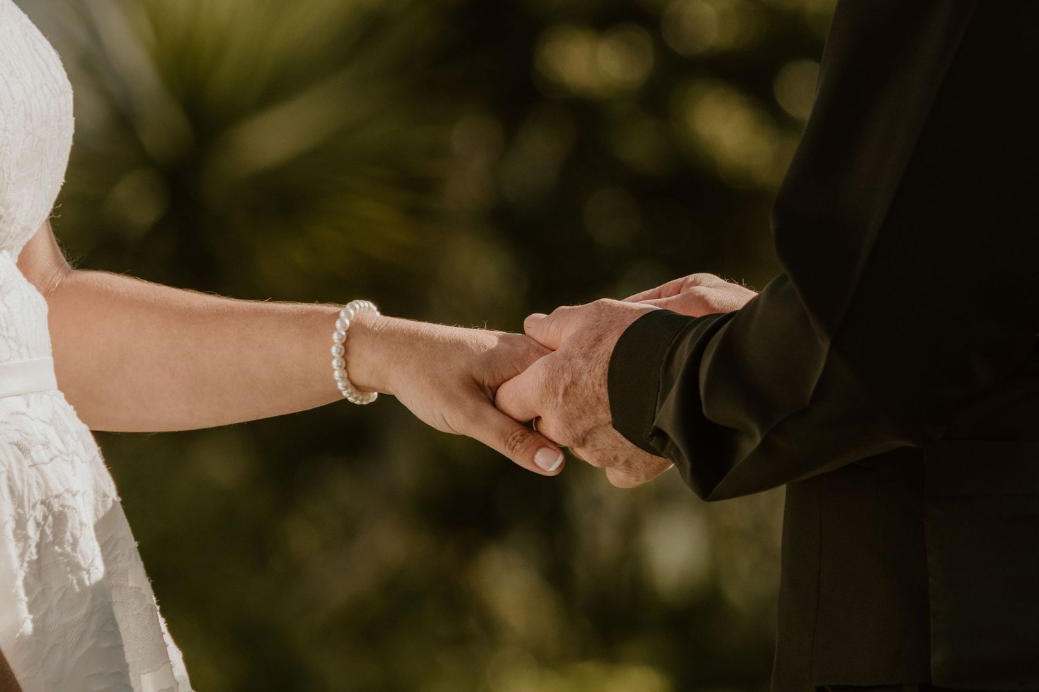 Close up of bride and groom holding hands with wedding attire and subtle jewelry visible in natural outdoor lighting.