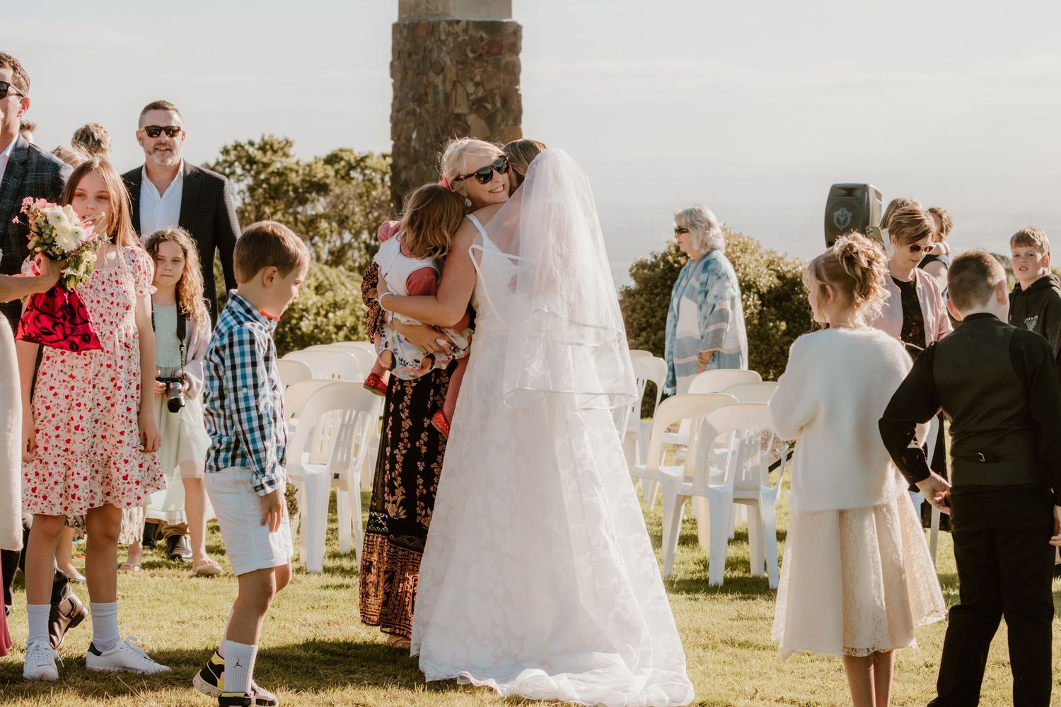 Wedding guests gather at an outdoor ceremony venue with white chairs set up on a sunny day overlooking scenic views.