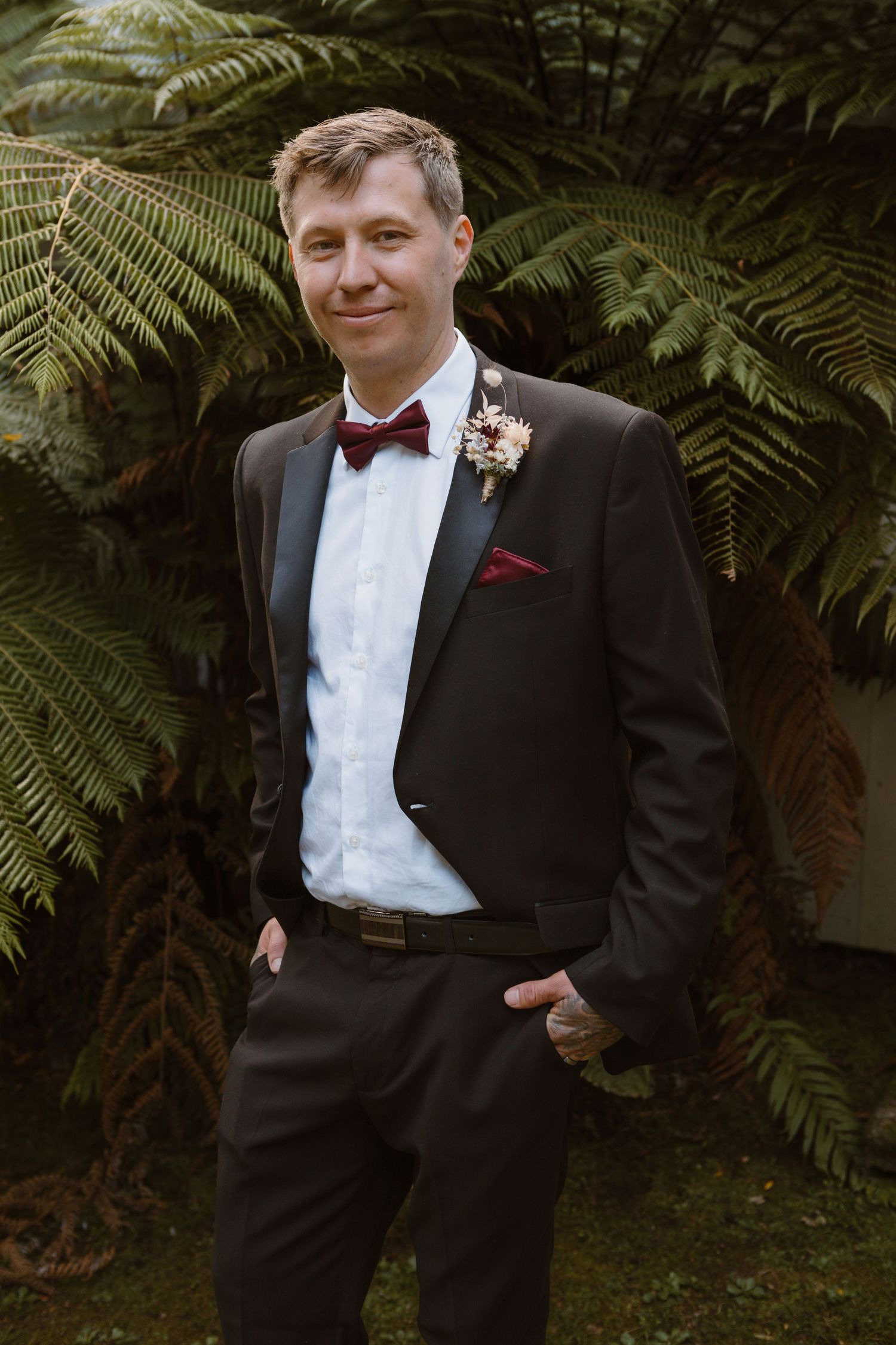 A groom in a black tuxedo with burgundy bow tie poses against lush fern foliage at an outdoor wedding.