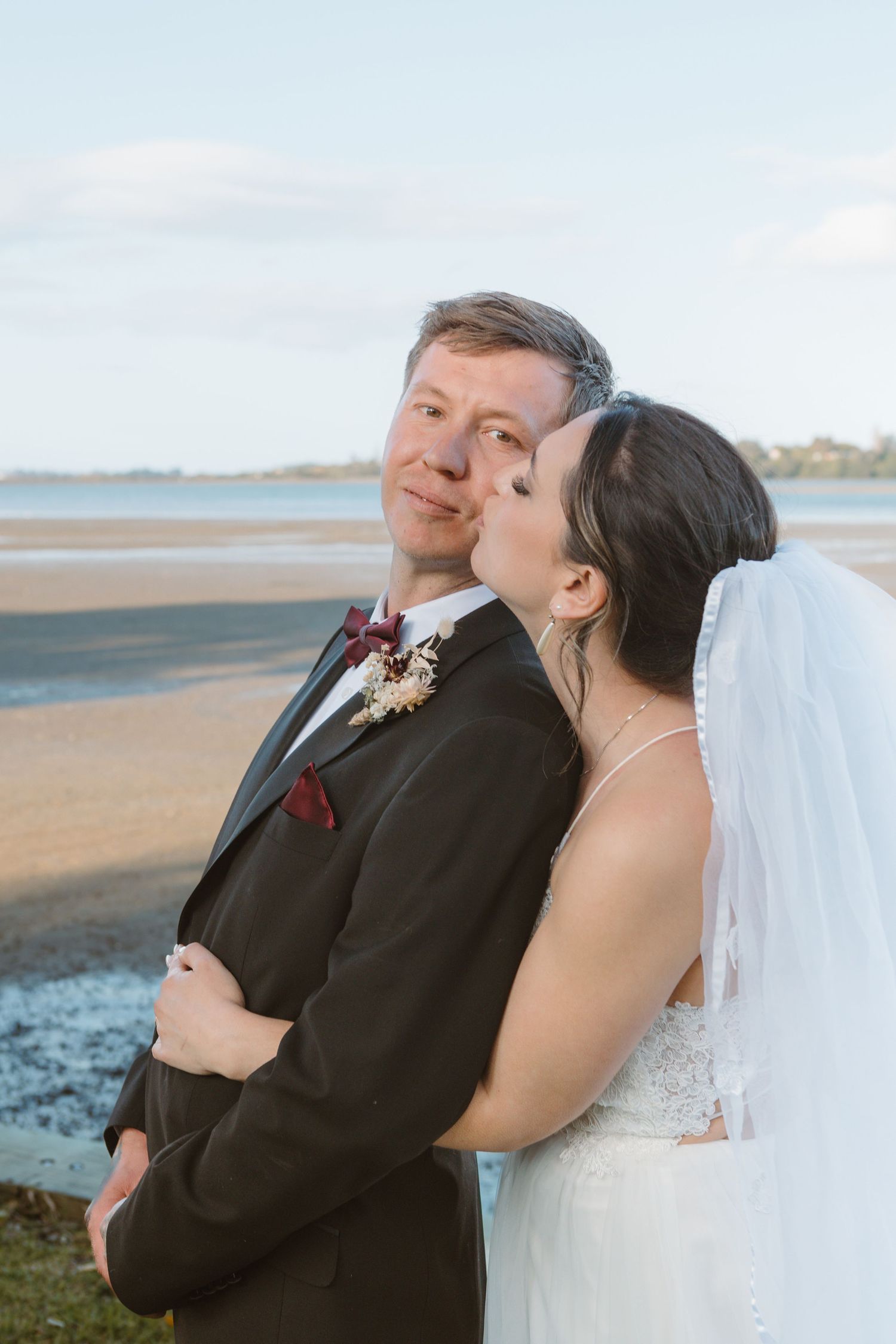 A couple shares a tender seaside moment in wedding attire at sunset on a beach.