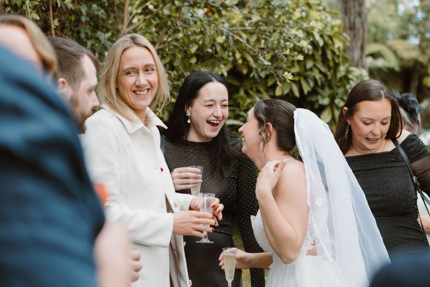 A joyful wedding celebration scene outdoors with guests laughing and socializing together while holding drinks.
