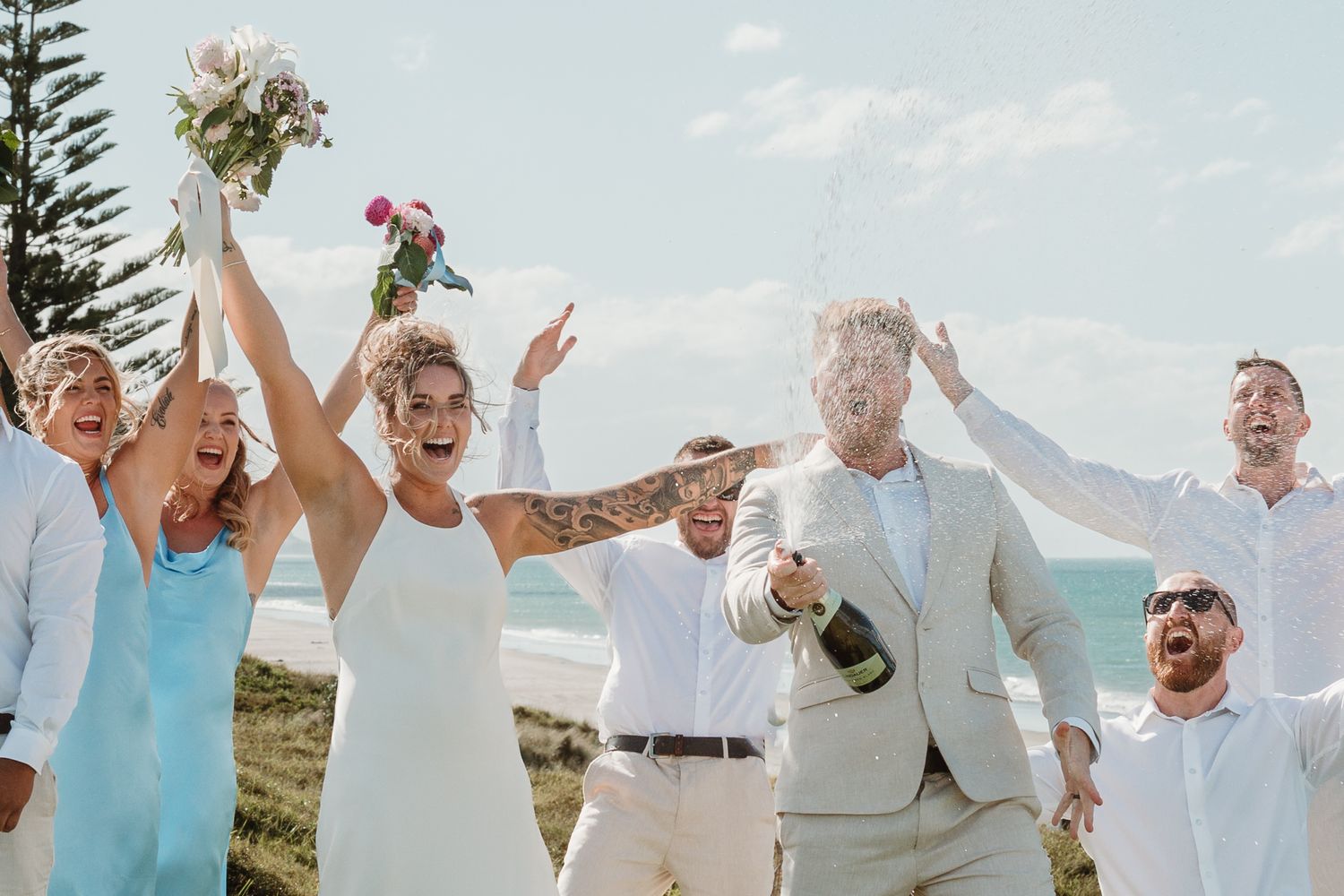 Joyful wedding party celebrating outdoors near the beach, with bride and groom popping champagne.
