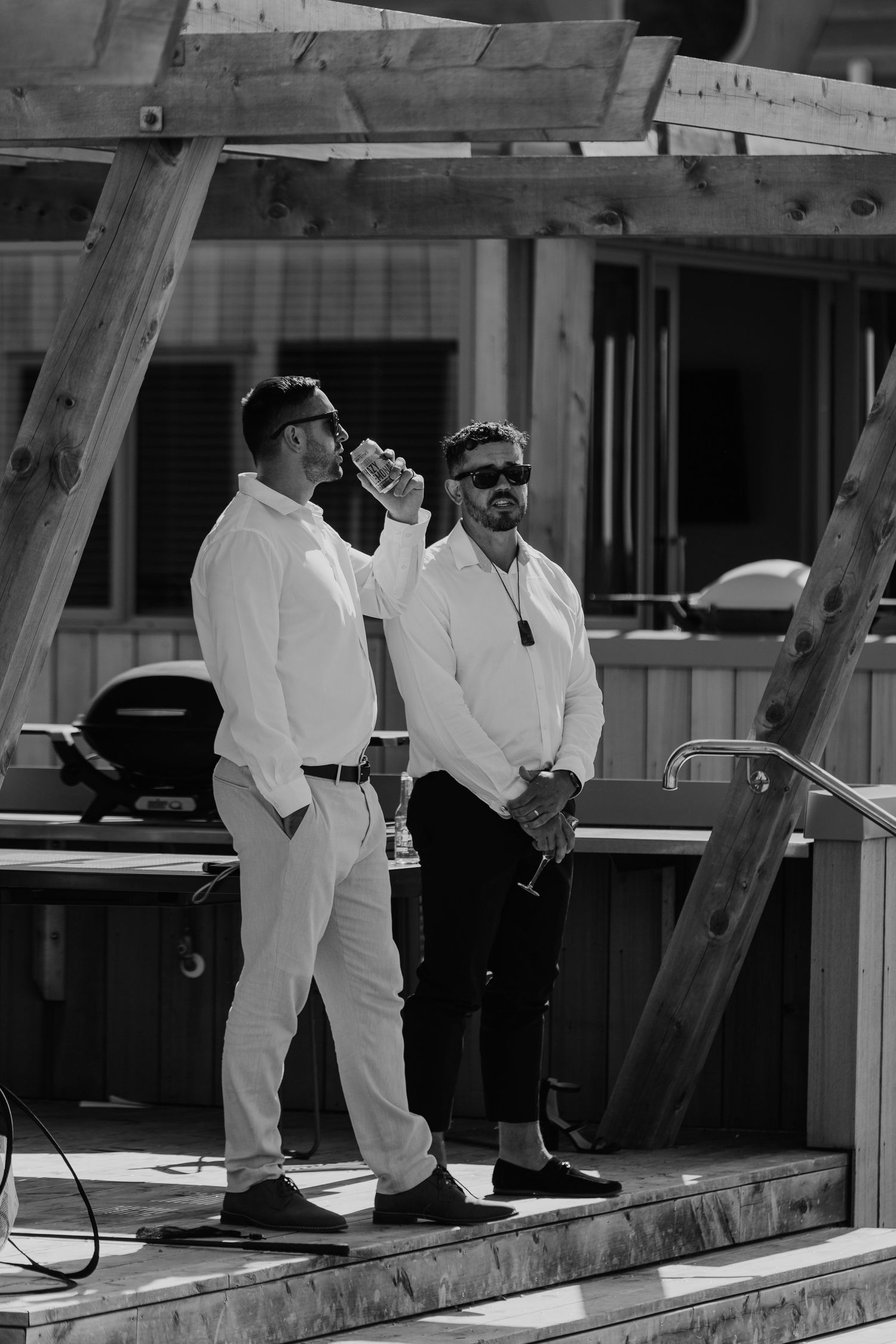 Two men in white shirts stand on a wooden deck, one drinking from a can, in a black and white outdoor photo.
