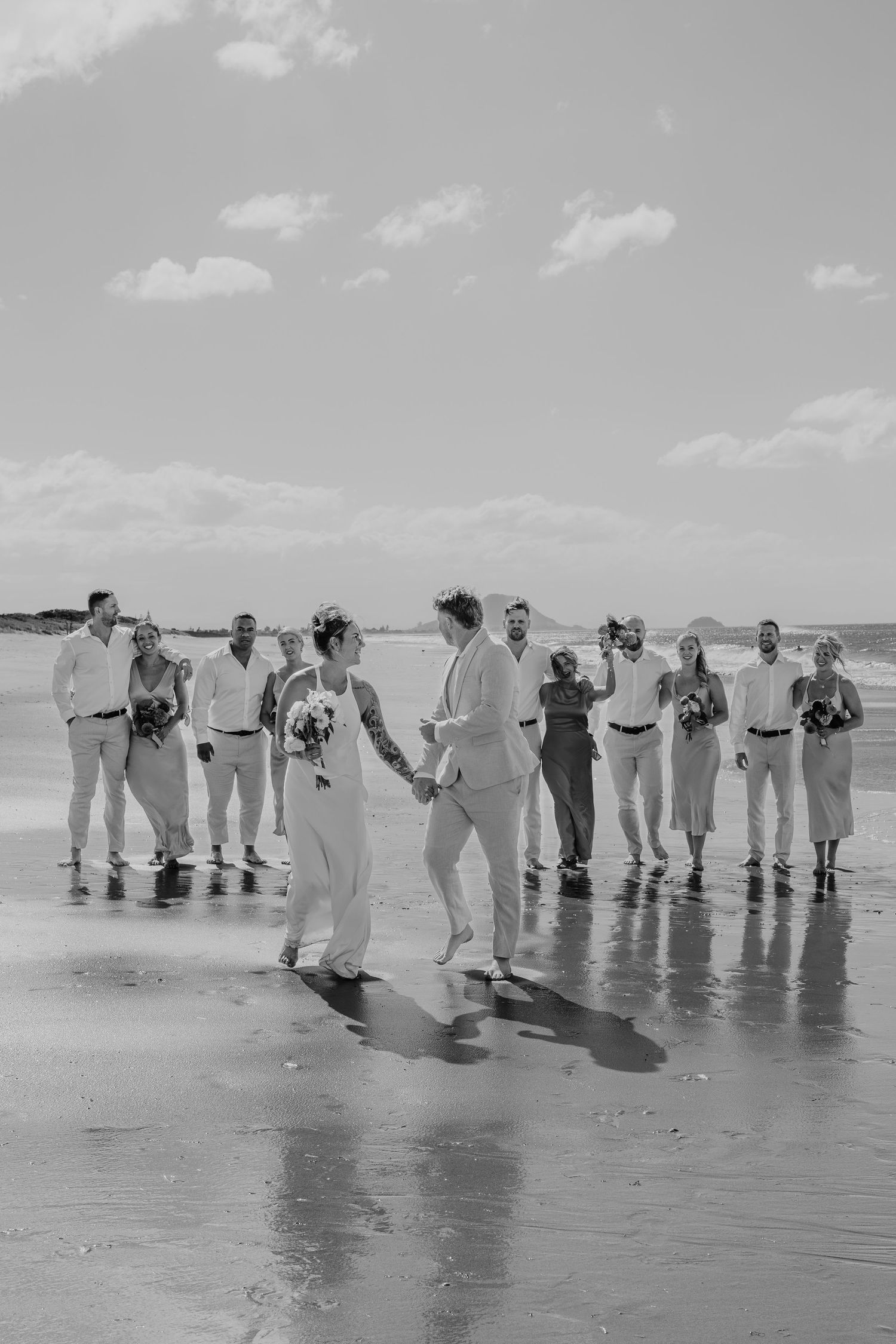 Black and white beach wedding photo with couple holding hands and bridal party celebrating on wet sand under cloudy sky.