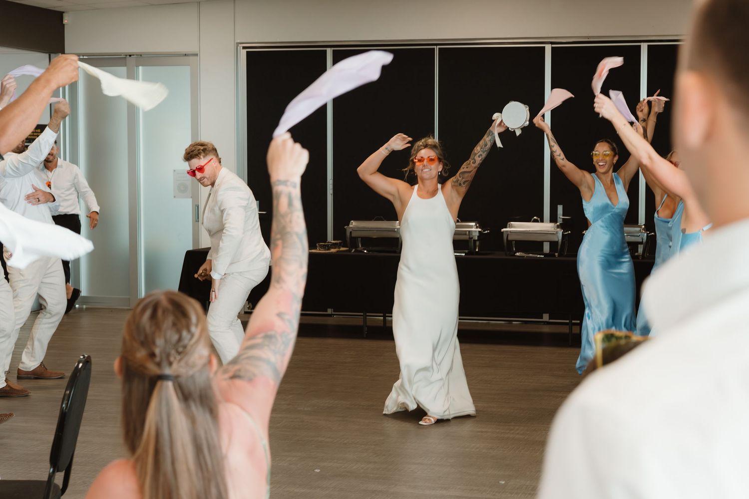 Bride in white gown waves napkin with guests during lively wedding reception celebration indoors.