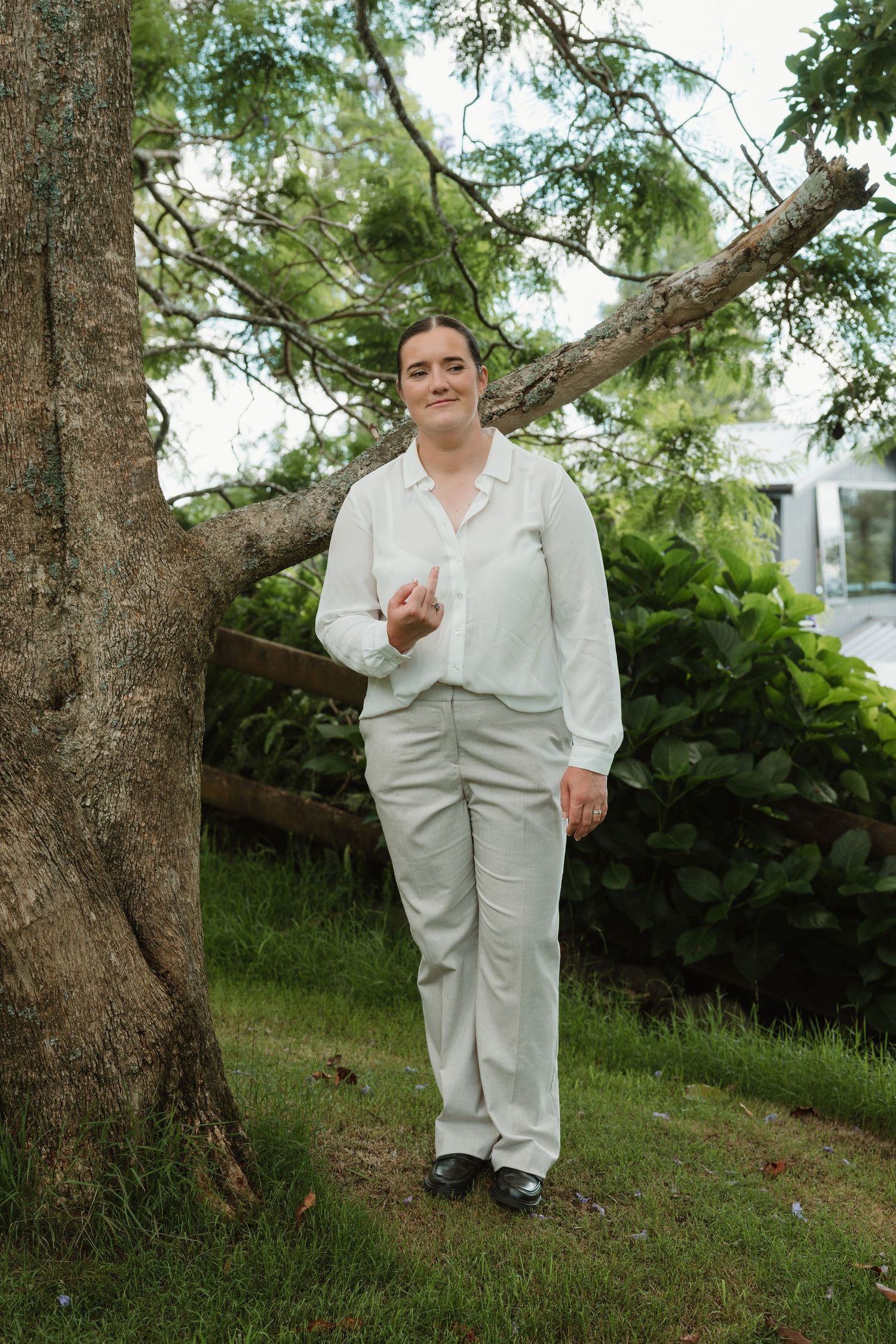 Person in white shirt and light pants standing by a tree in a lush green garden, smiling warmly.