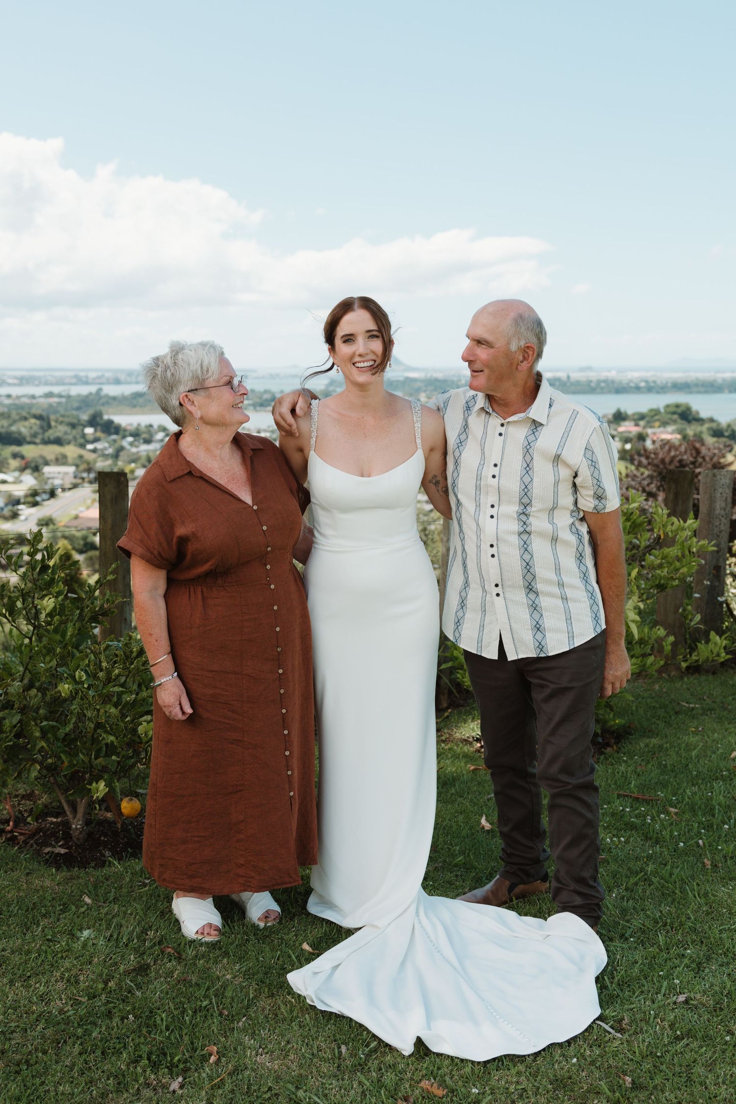 Bride in white gown poses with older couple outdoors on green lawn with scenic water view on wedding day.