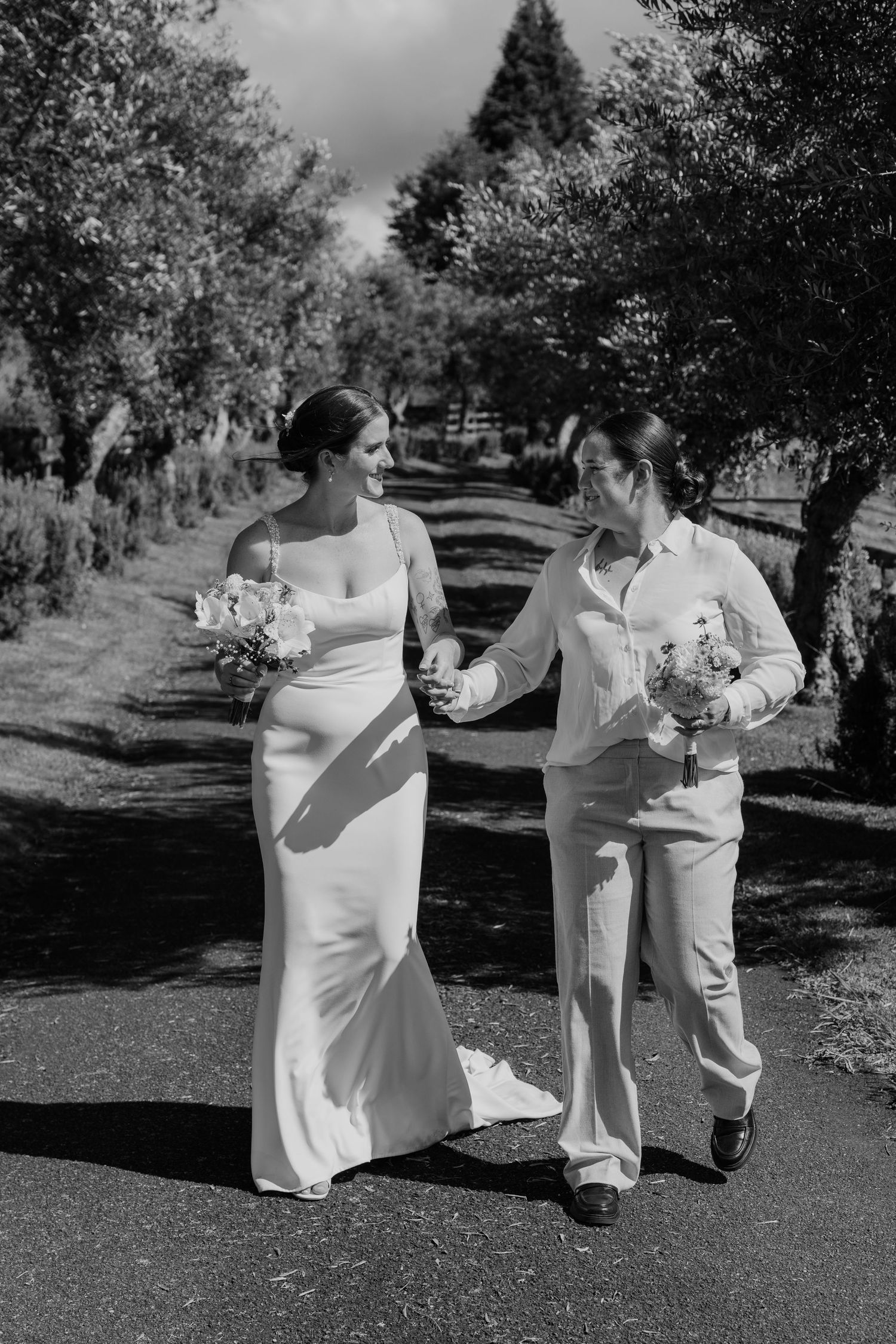 Two brides walk hand-in-hand down a tree-lined path, one in a gown and one in a suit, holding bouquets.