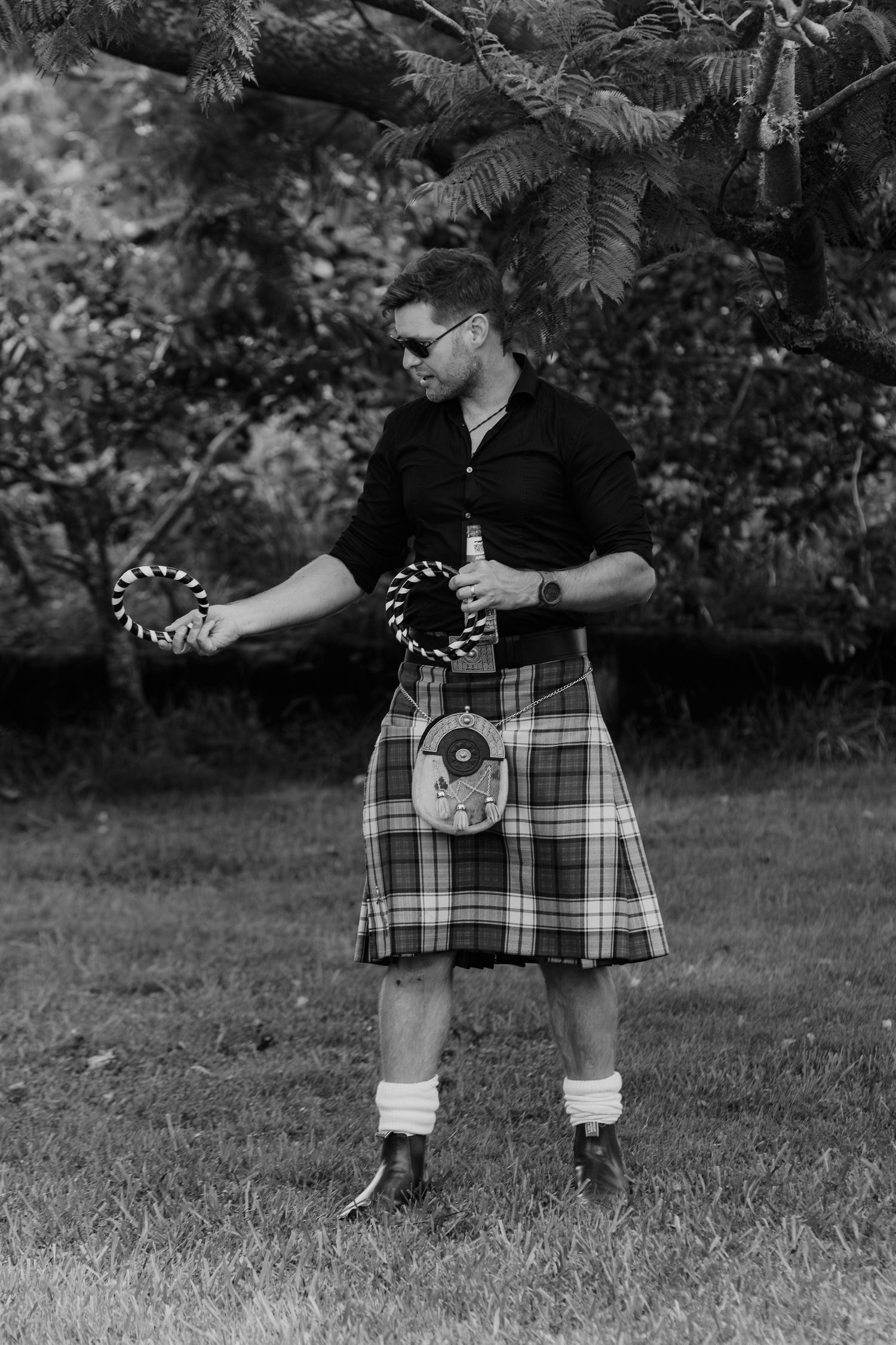 Man in traditional Scottish kilt and sporran holds tambourines outdoors in black and white photo.