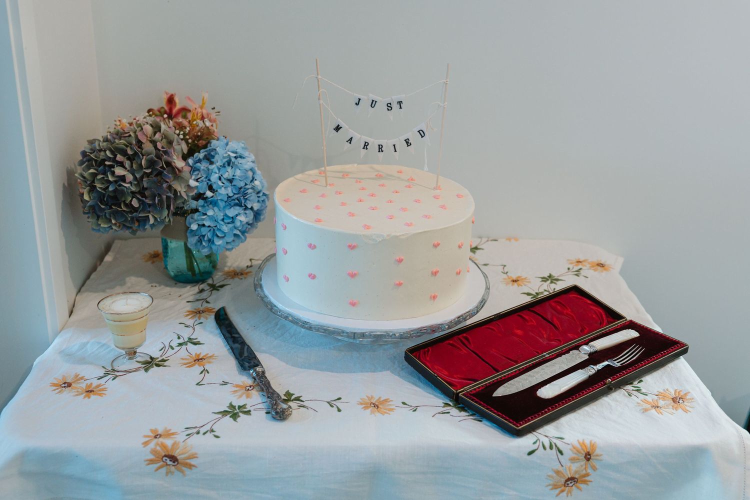White wedding cake with pink floral details and 'Just Married' banner on a floral tablecloth with cake knives.