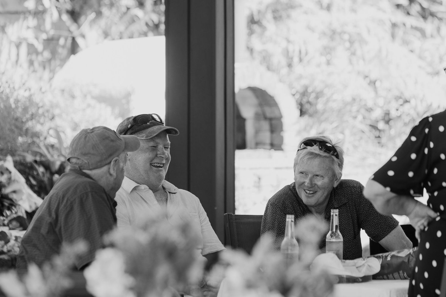 Three people laughing together at an outdoor table, sharing drinks in a candid black and white photo.