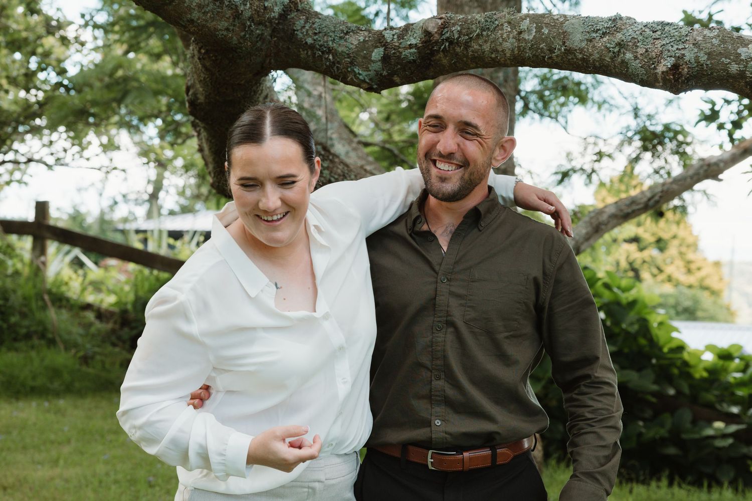 A smiling couple poses outdoors under a tree, woman in white shirt and man in olive green shirt.
