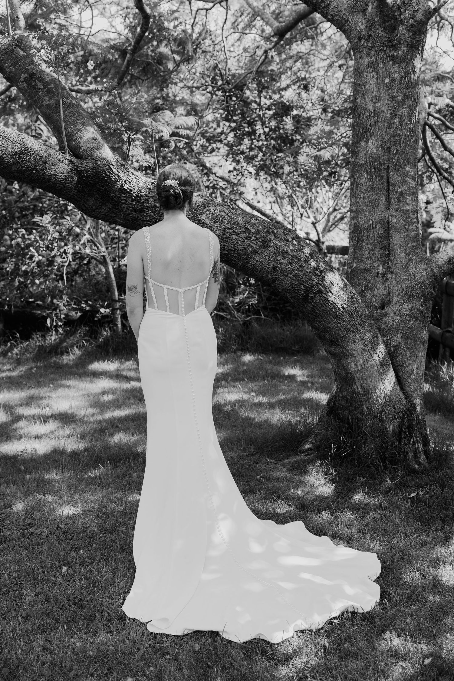 Black and white photo of a bride in a backless wedding gown with a train, standing before a large tree outdoors.