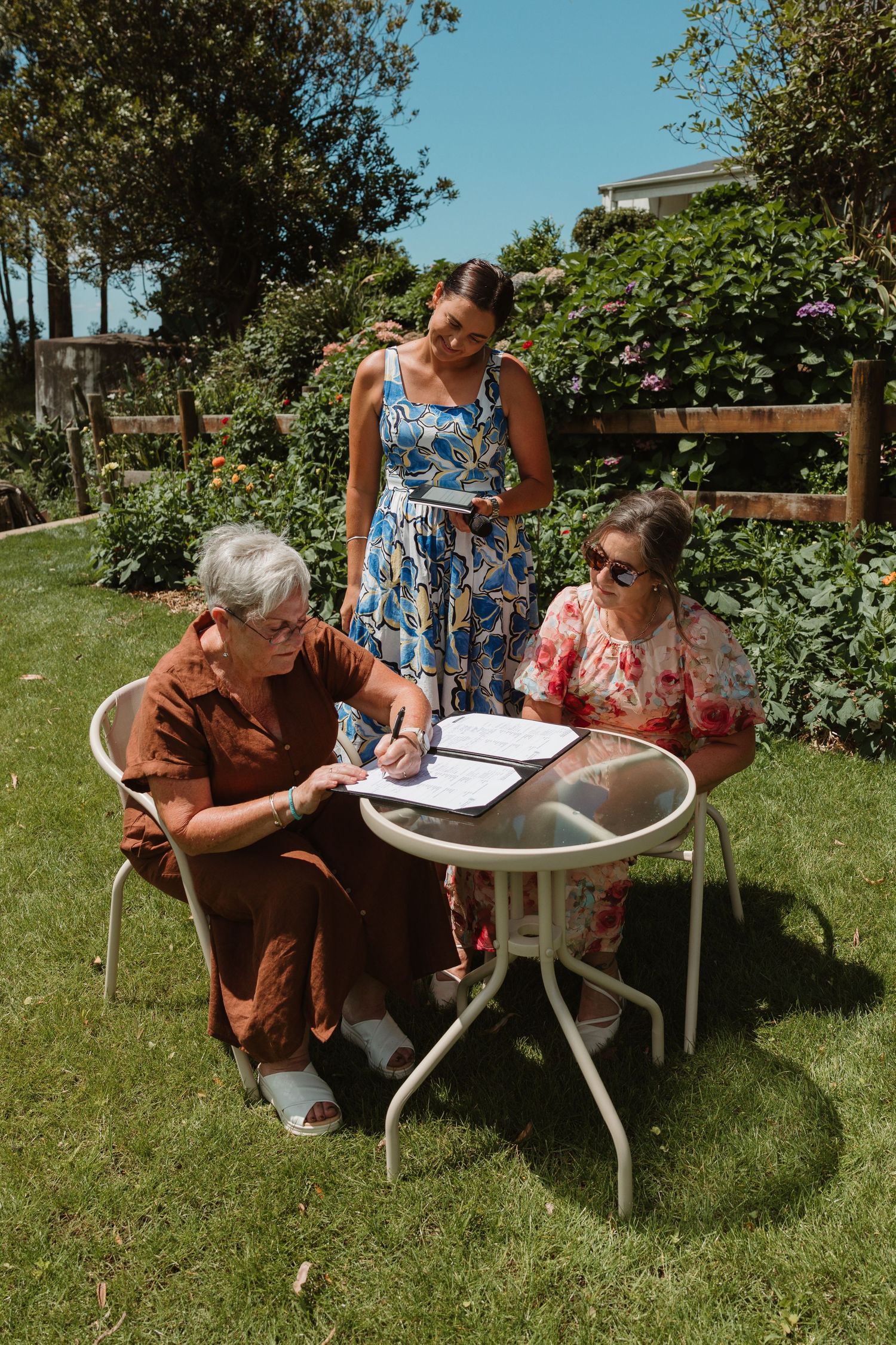 Three women reviewing and signing documents at an outdoor garden table on a sunny day.