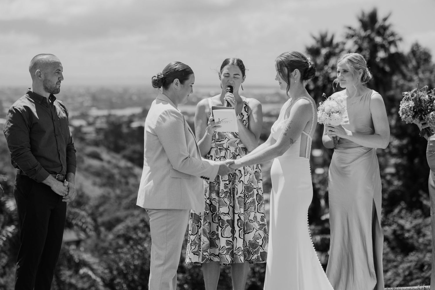 Black and white wedding ceremony photo of two brides holding hands with officiant and bridesmaid outdoors.