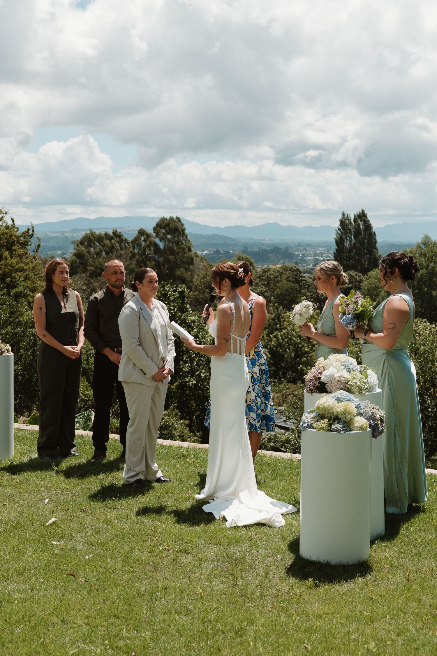 Outdoor wedding ceremony on a hilltop with bride, groom, bridesmaids, and guests surrounded by scenic mountain views.