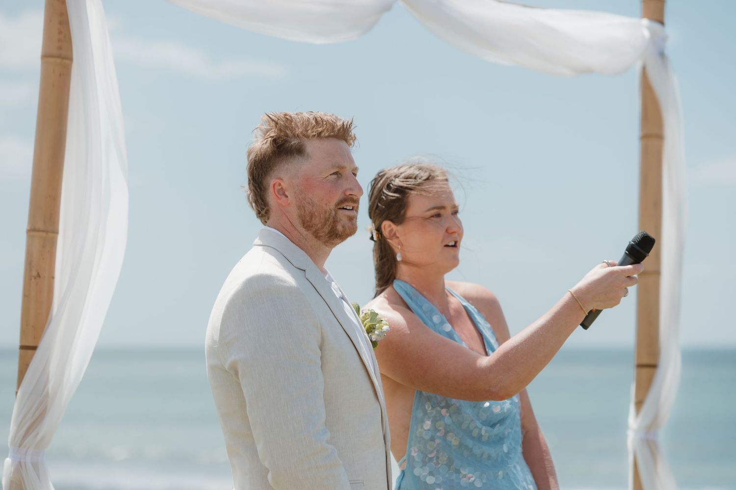 Couple stands under bamboo arch at beach wedding ceremony, woman in blue floral dress holds microphone.