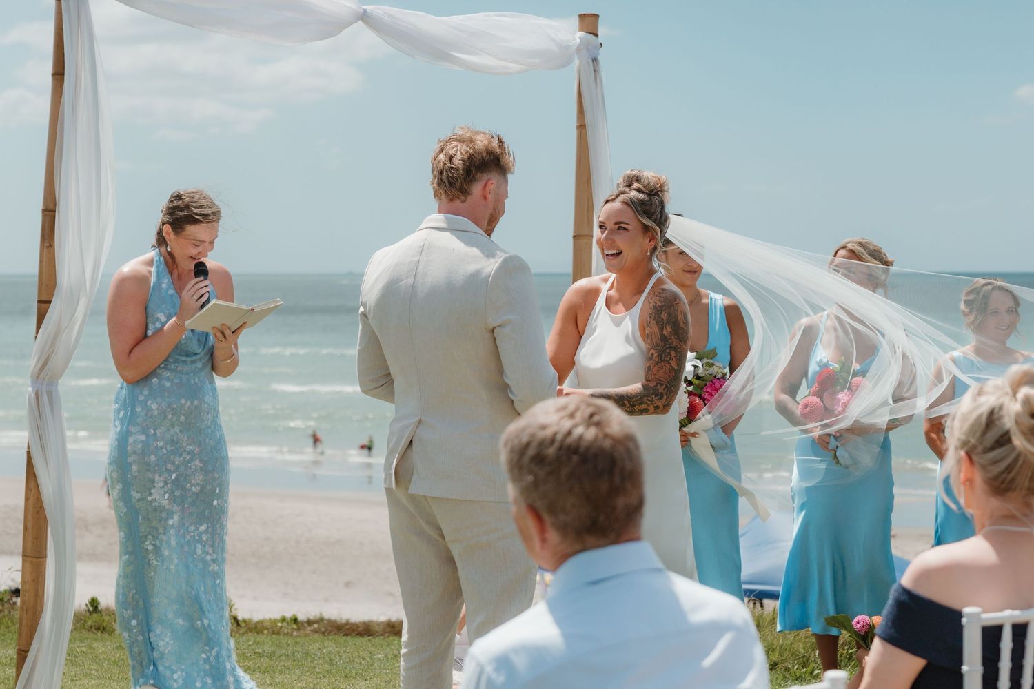Bride and groom exchange vows at a beachside wedding ceremony with bridesmaids in blue dresses on a sunny day.