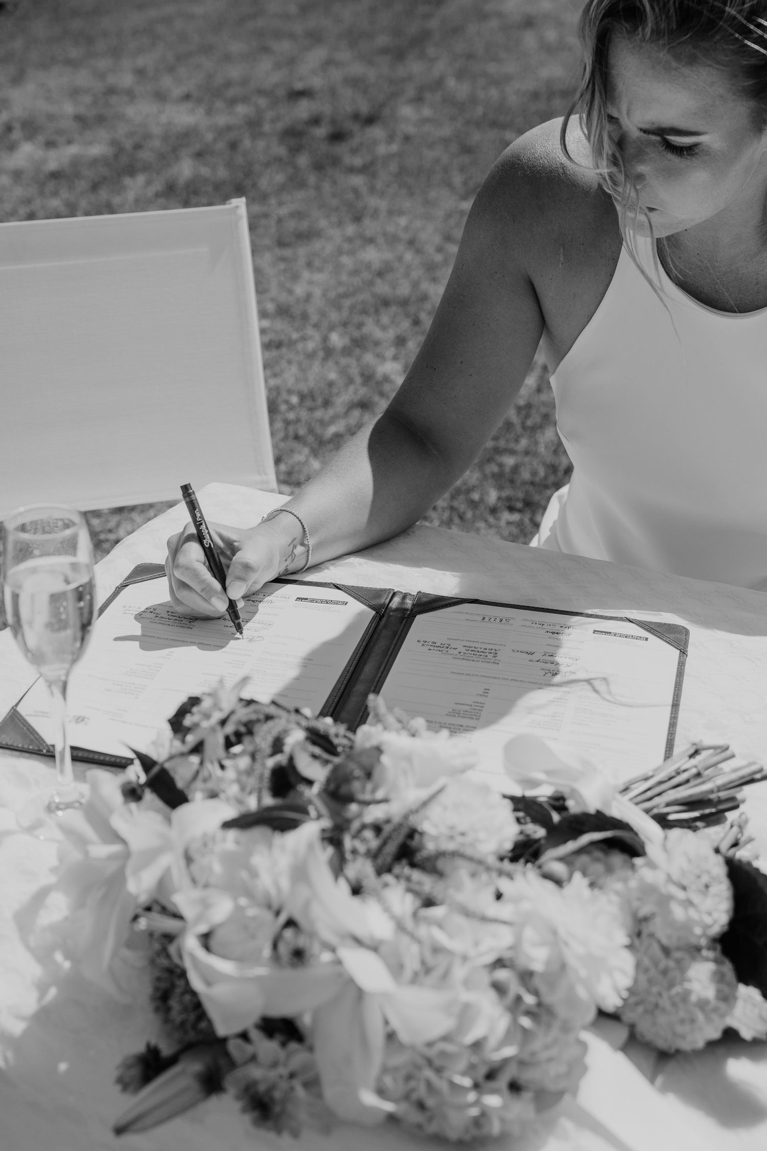 Bride signing wedding registry documents at outdoor table with floral bouquet and champagne glass in black and white photo.