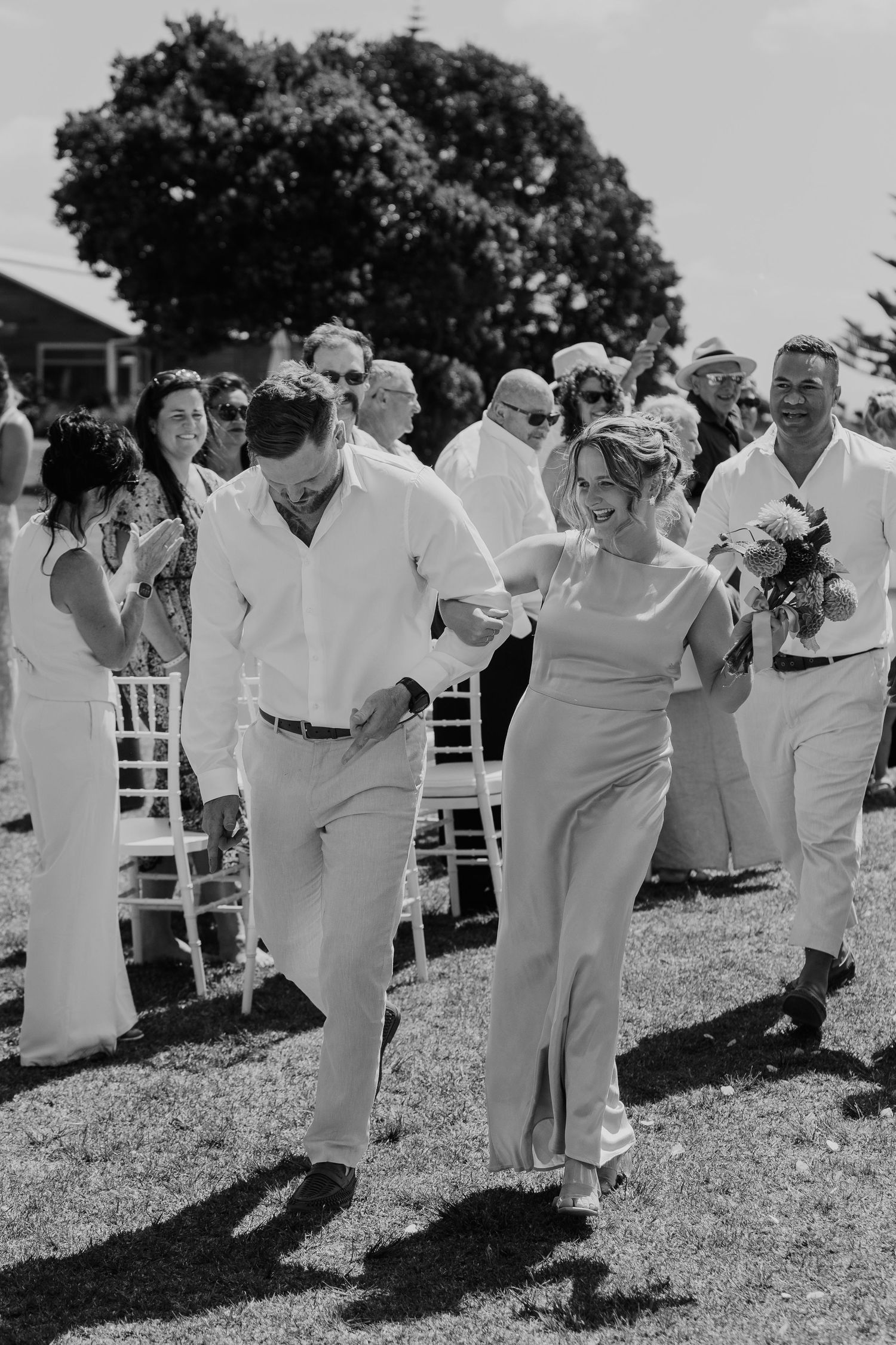 Black and white photo of a joyful couple walking down the aisle at an outdoor wedding ceremony with guests applauding.