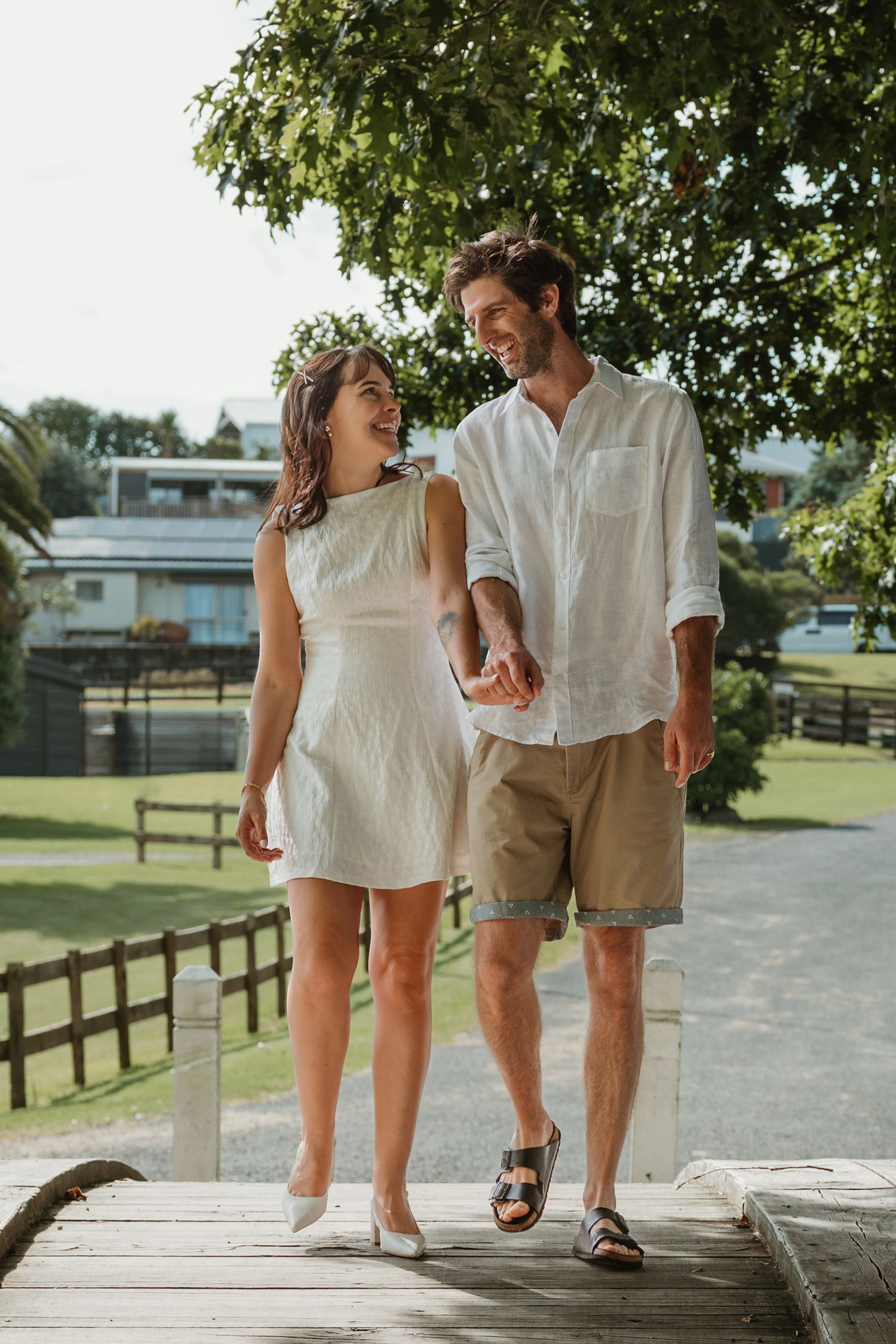 A smiling couple holds hands while walking on a wooden deck outdoors, dressed in casual summer attire.