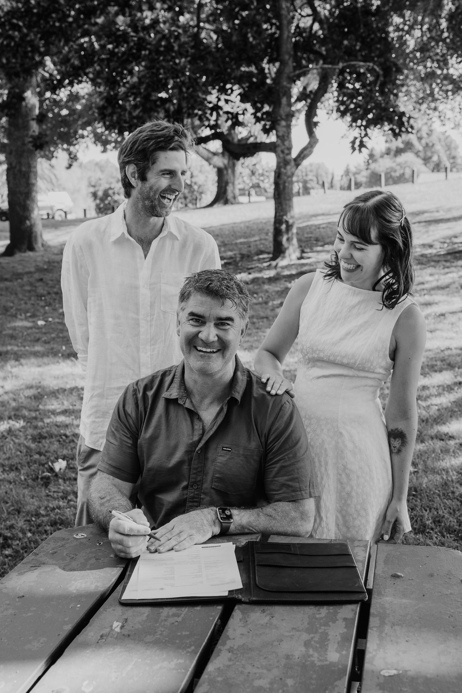 Black and white photo of a couple signing wedding documents at a picnic table outdoors, officiant seated smiling.