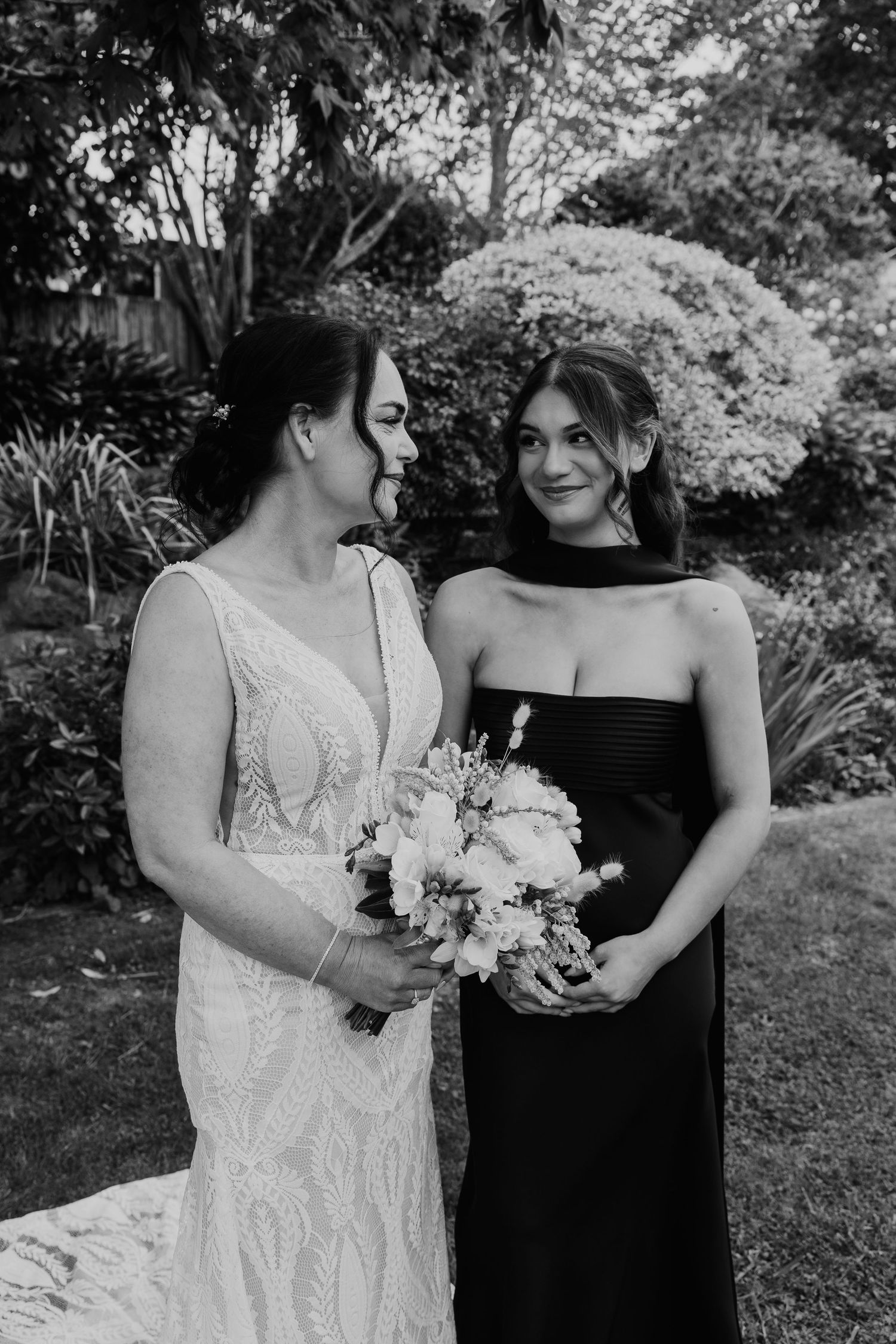 Two people in contrasting black and white outfits stand together holding floral bouquets in an outdoor garden setting.