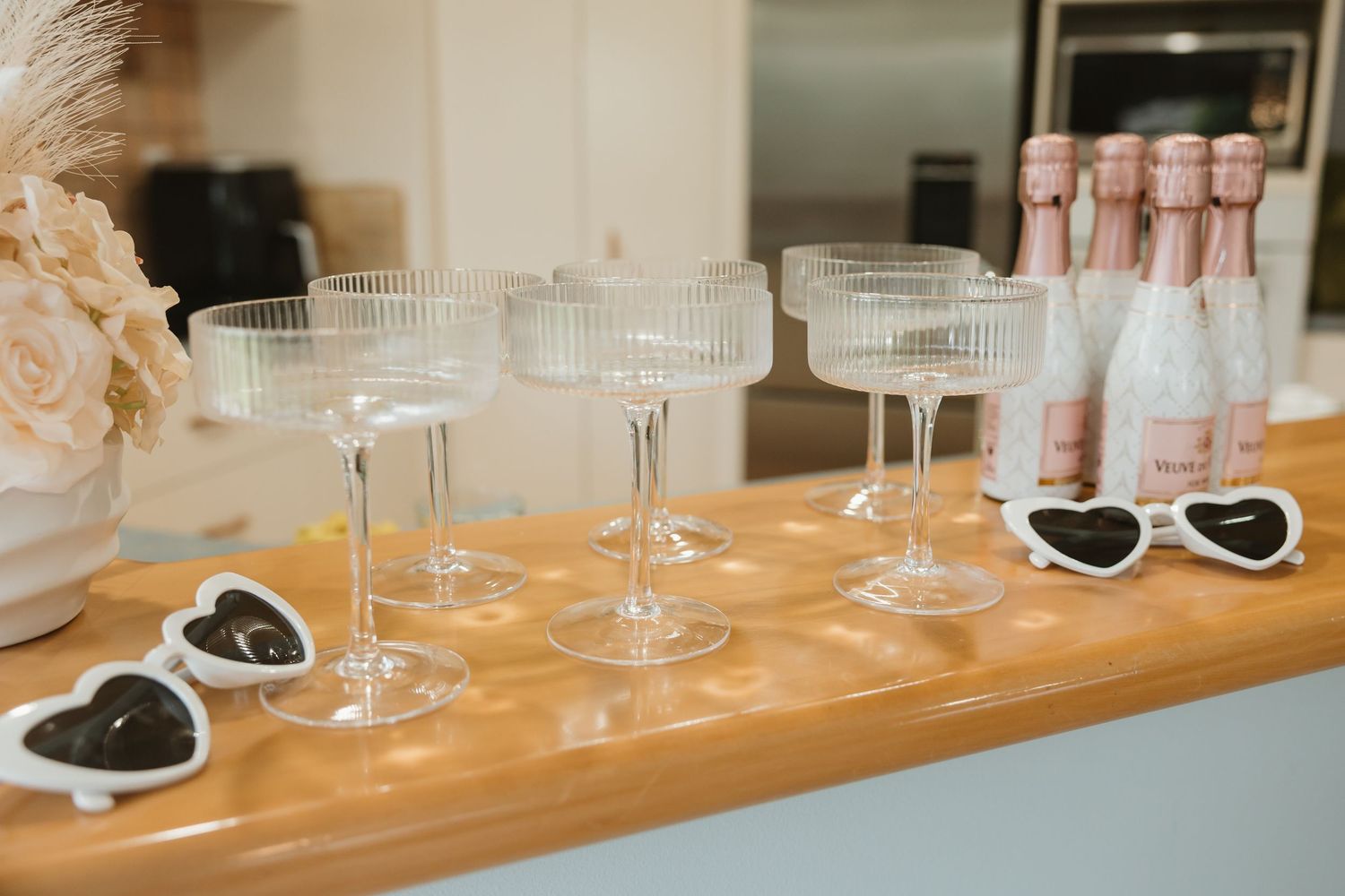 Vintage-style champagne coupes and heart-shaped sunglasses arranged on a wooden table with pink roses and bottles.