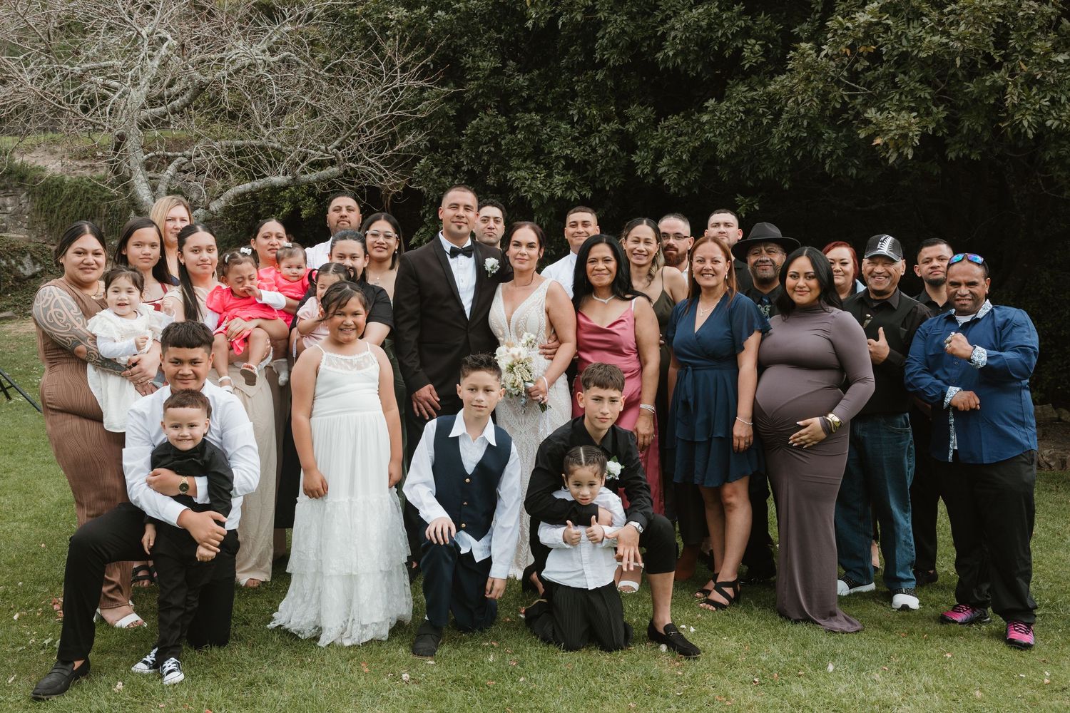 A large group of wedding guests pose together outdoors for a series of formal group photos in a garden setting.