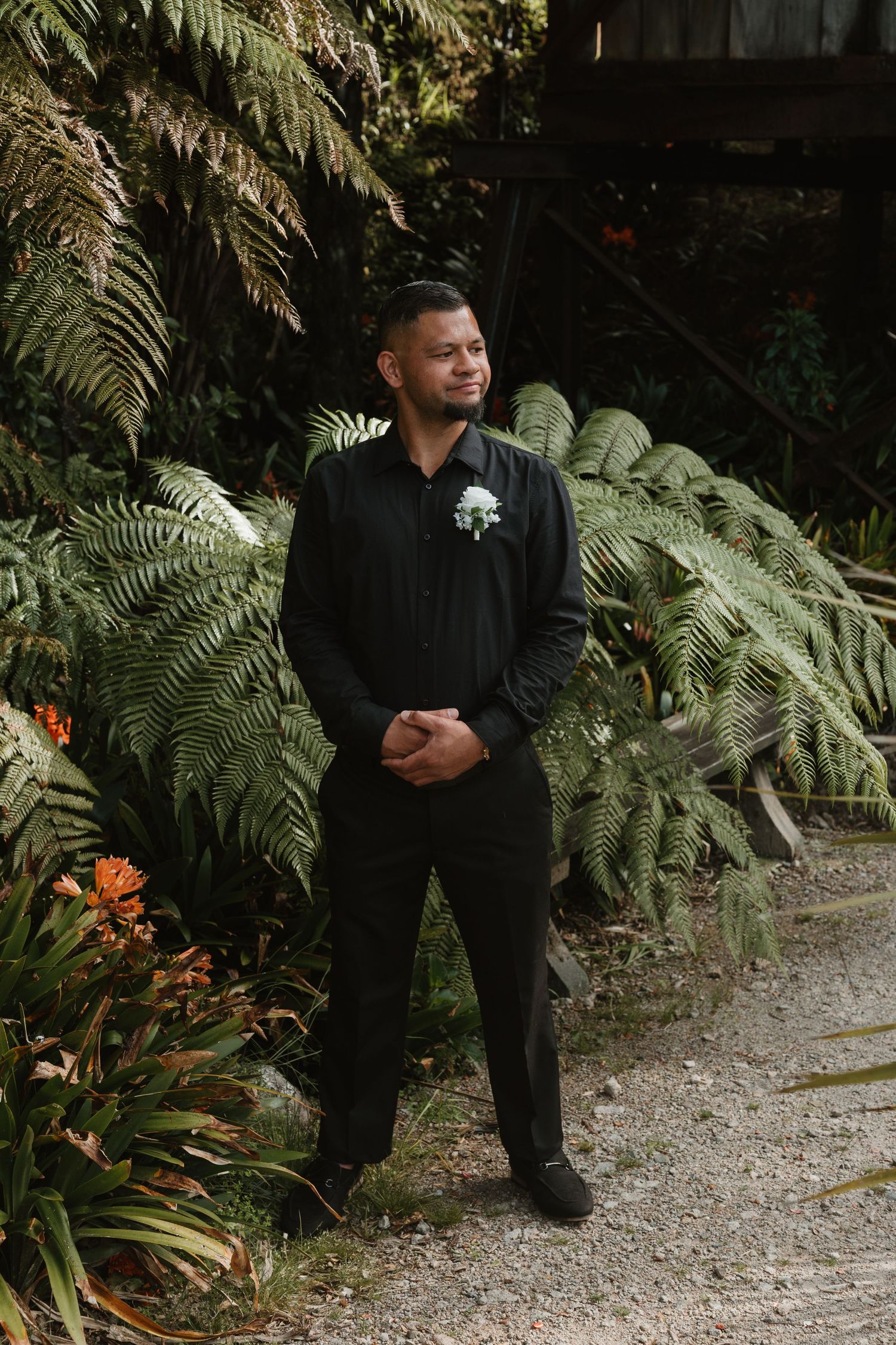 A person in formal black attire stands on a garden path surrounded by lush tropical ferns and greenery.