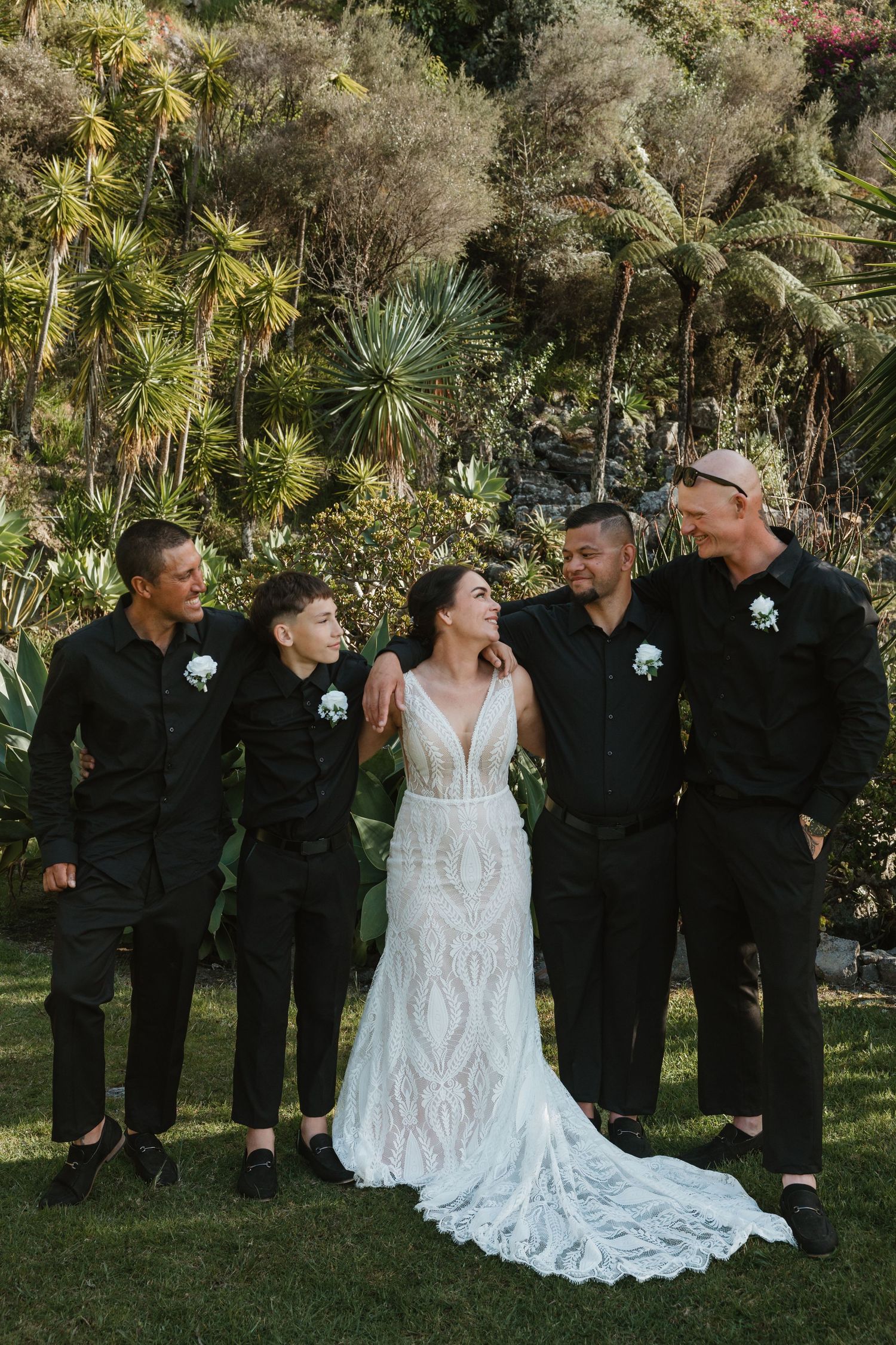 Wedding party in black attire stands with bride in white gown against lush garden backdrop.